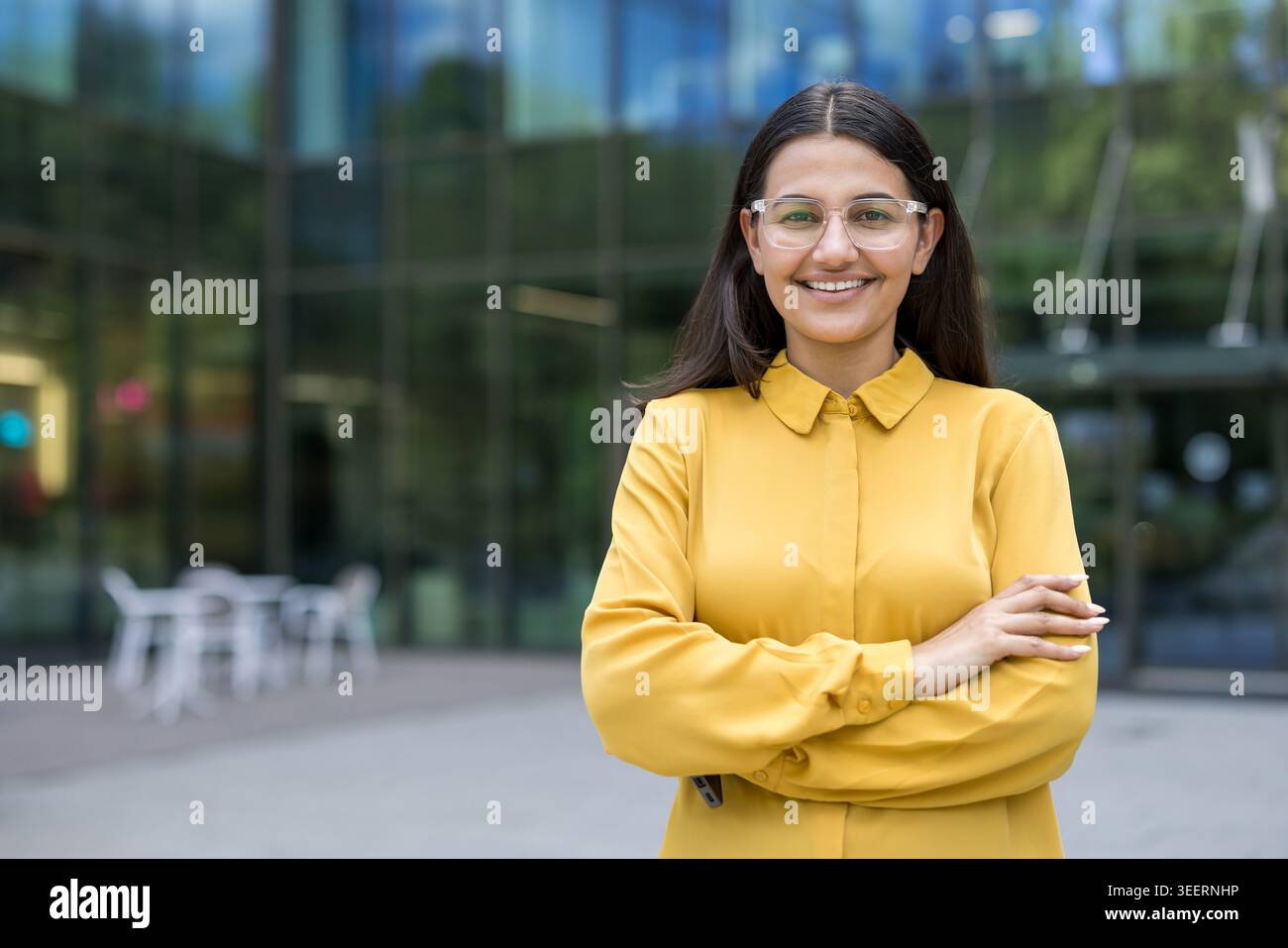 Jeune femme professionnelle croisant les bras, portant des lunettes transparentes et une chemise jaune, souriant à l'extérieur d'un immeuble de bureaux en verre moderne, représentant confiance et succès Banque D'Images