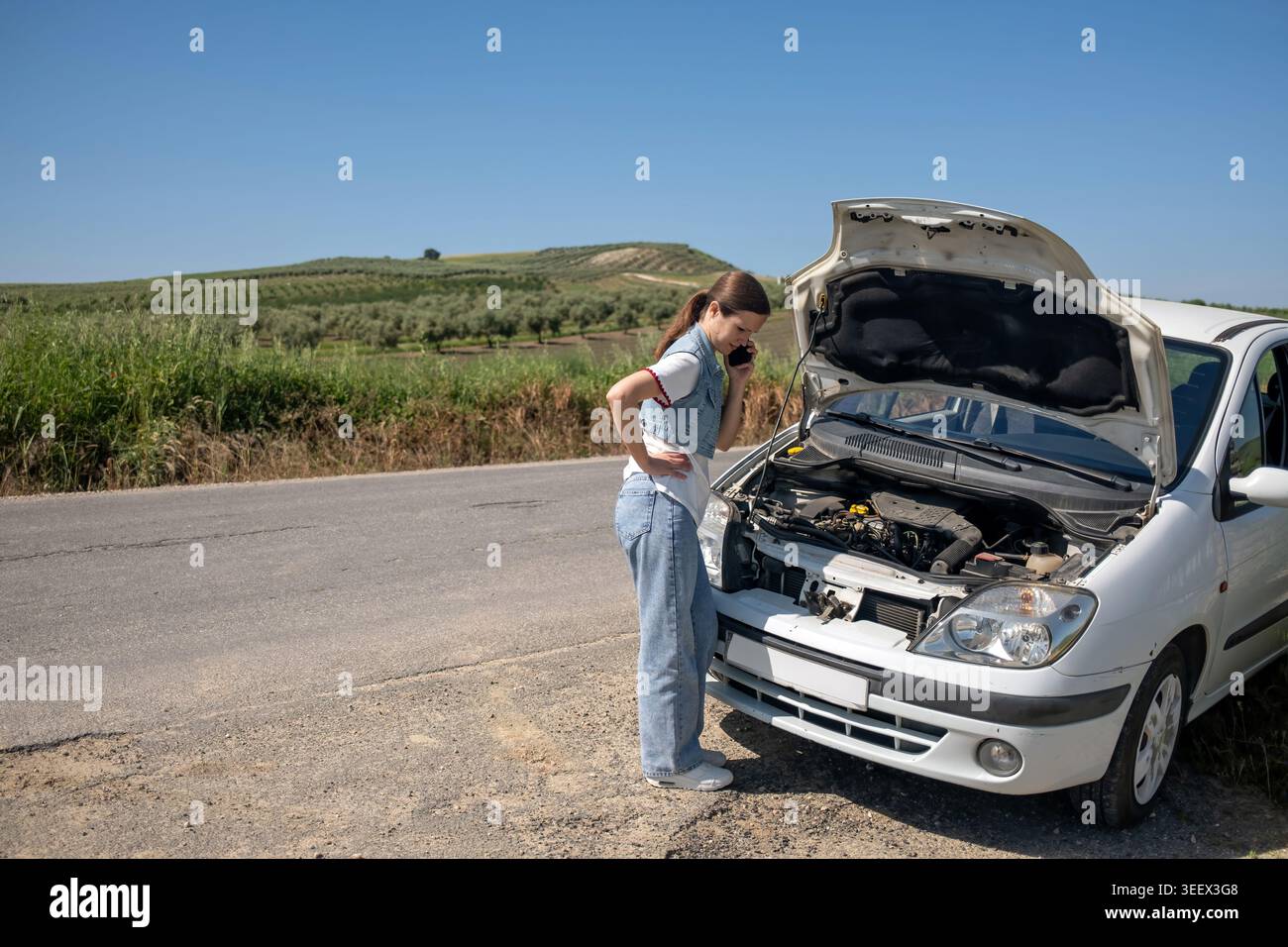 Femme debout à côté d'un véhicule cassé avec le capot ouvert, faisant un appel téléphonique pour l'assistance routière sur une route rurale Banque D'Images