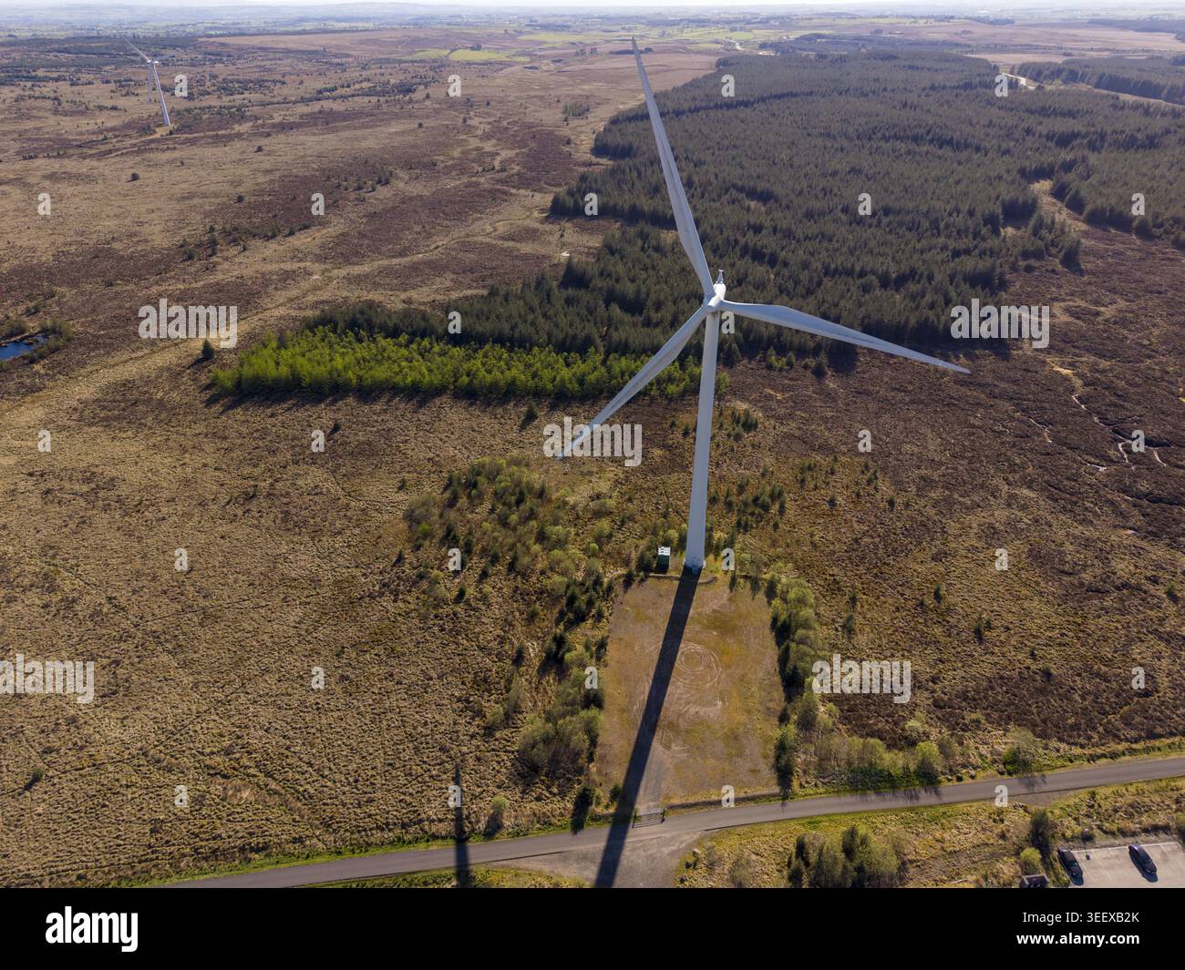 Vue aérienne d'une grande éolienne blanche debout dans un vaste paysage rural avec des forêts denses et des champs ouverts sous un ciel dégagé. Banque D'Images