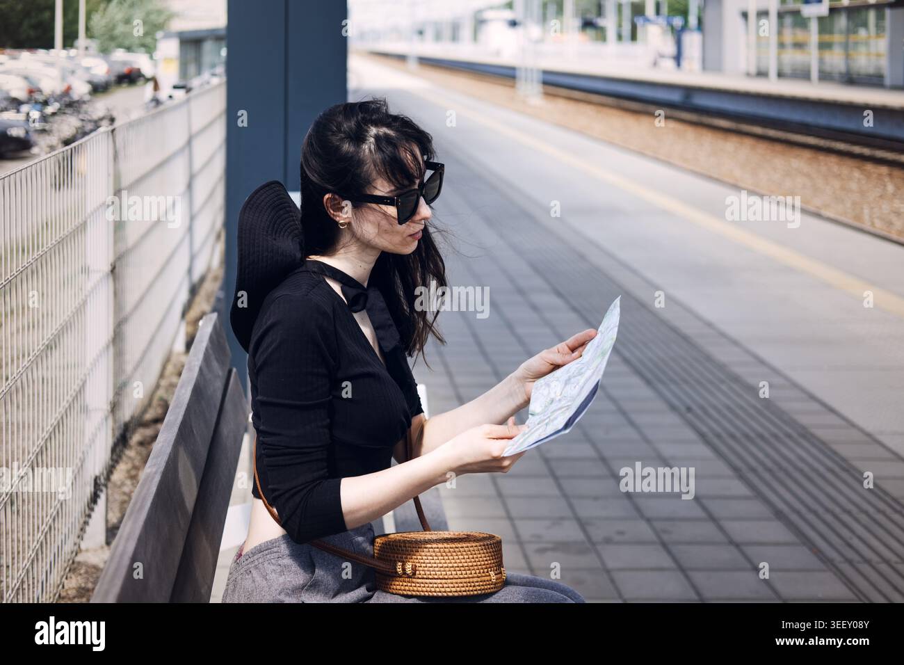 La jeune femme est assise sur le banc au quai de chemin de fer tout en étudiant la carte papier. Voyage conscient, vacances de détox numérique, style de voyage nostalgique. Banque D'Images