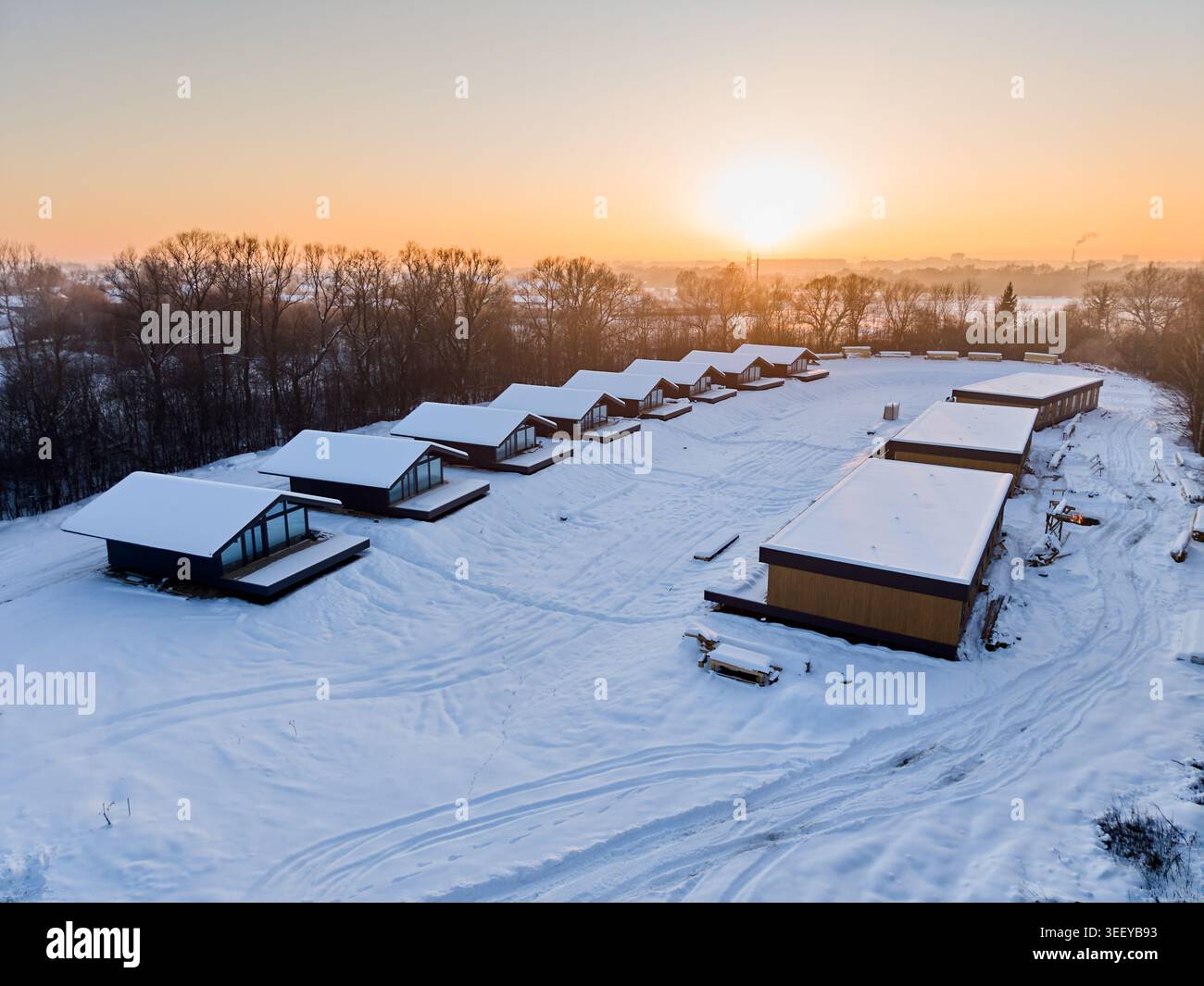 Rangée de cabines à ossature en bois modernes dans un paysage enneigé au coucher du soleil, vue aérienne du développement de la station éco avec des maisons minimalistes et une lumière chaude créant une atmosphère hivernale paisible Banque D'Images