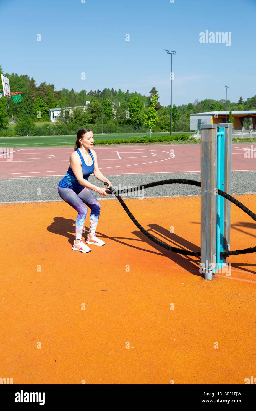 Femme faisant l'entraînement de corde de combat sur la salle de gym extérieure dans le parc urbain ensoleillé. Entraînement fitness puissant et authentique sur surface sportive orange. Banque D'Images