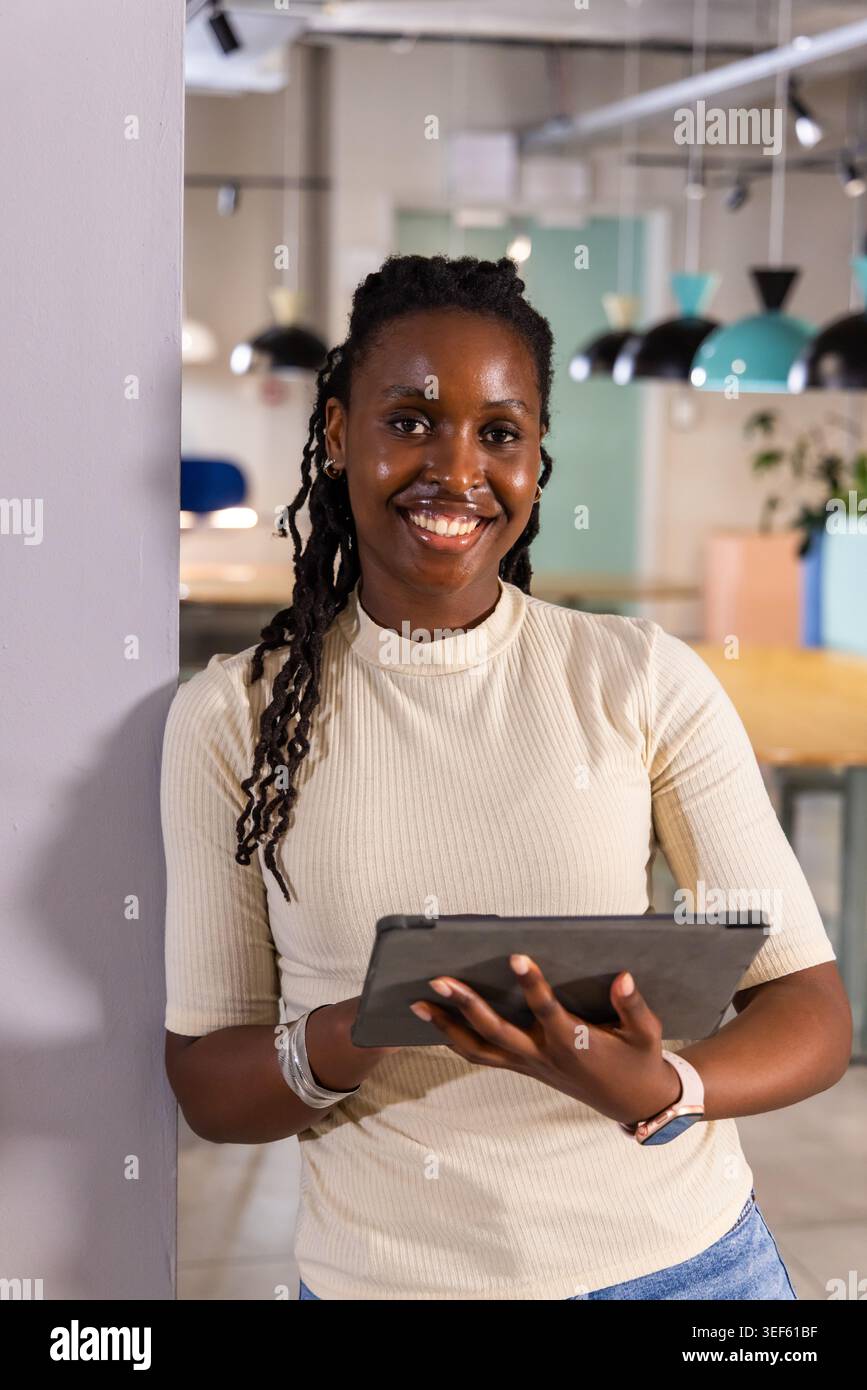 Femme afro-américaine appuyée contre la colonne dans le bureau, souriante et tenant la tablette avec smartwatch Banque D'Images