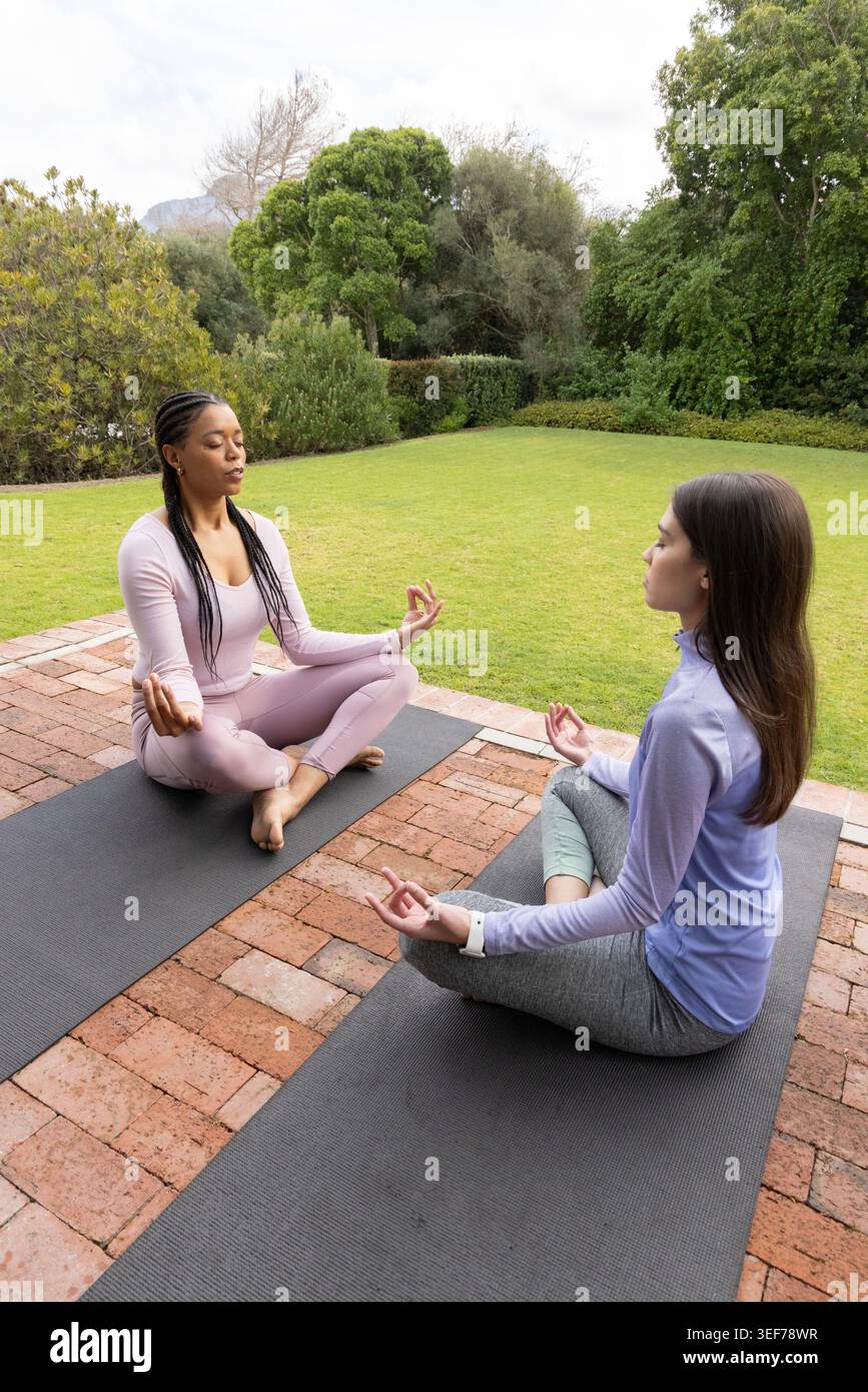 Diverses femelles méditant sur des tapis de yoga sombres sur le patio en briques dans le jardin en vêtements de sport avec smartwatch Banque D'Images