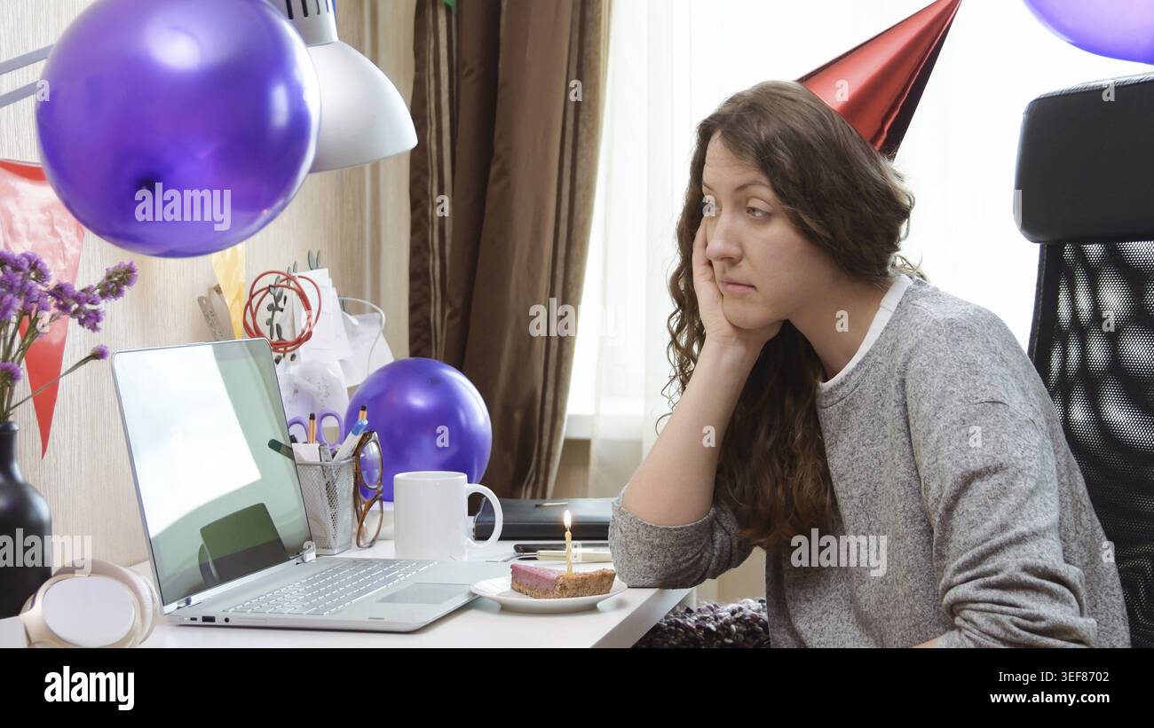 Triste femme célibataire célébrant son anniversaire par la fête virtuelle d'appel vidéo. Assis seul avec un ordinateur portable et gâteau avec une bougie. Lieu de travail authentique. Coron Banque D'Images