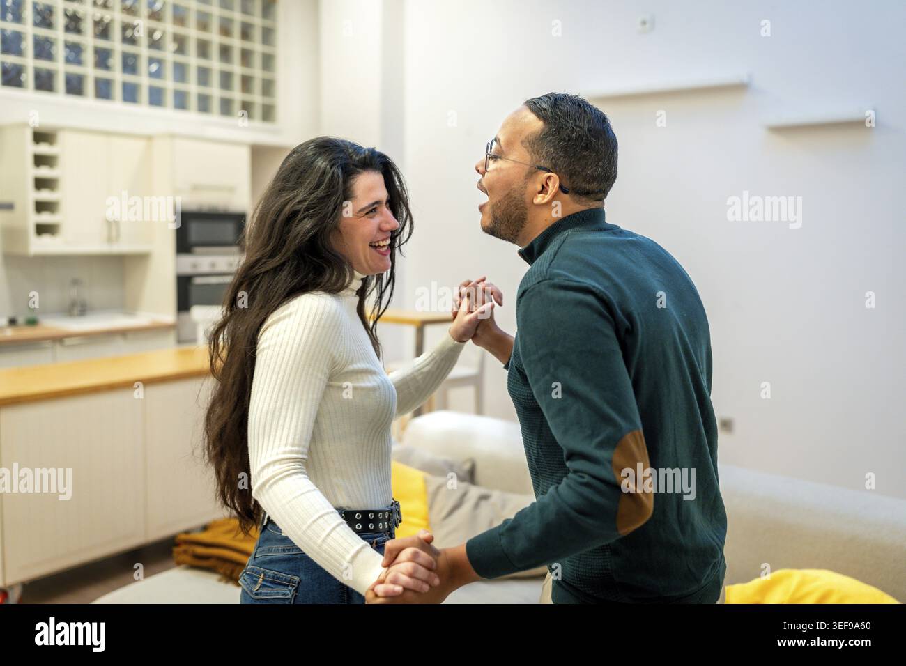 Heureux couple diversifié tenant la main et dansant intimement dans un salon et une cuisine ouverts modernes, profitant d'un joyeux moment de connexion et Mo Banque D'Images