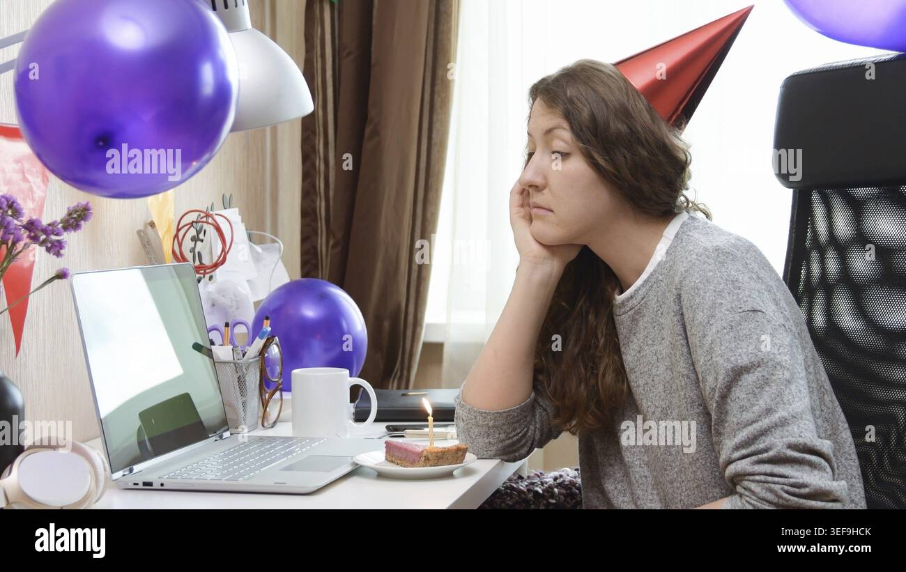 Triste femme célibataire célébrant son anniversaire par la fête virtuelle d'appel vidéo. Assis seul avec un ordinateur portable et gâteau avec une bougie. Lieu de travail authentique. Coron Banque D'Images