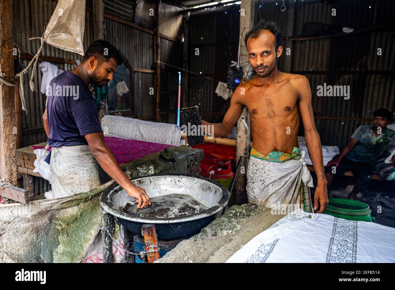 Un homme teint des vêtements de batik dans un atelier tandis qu'un autre prépare des matériaux au Bangladesh Banque D'Images
