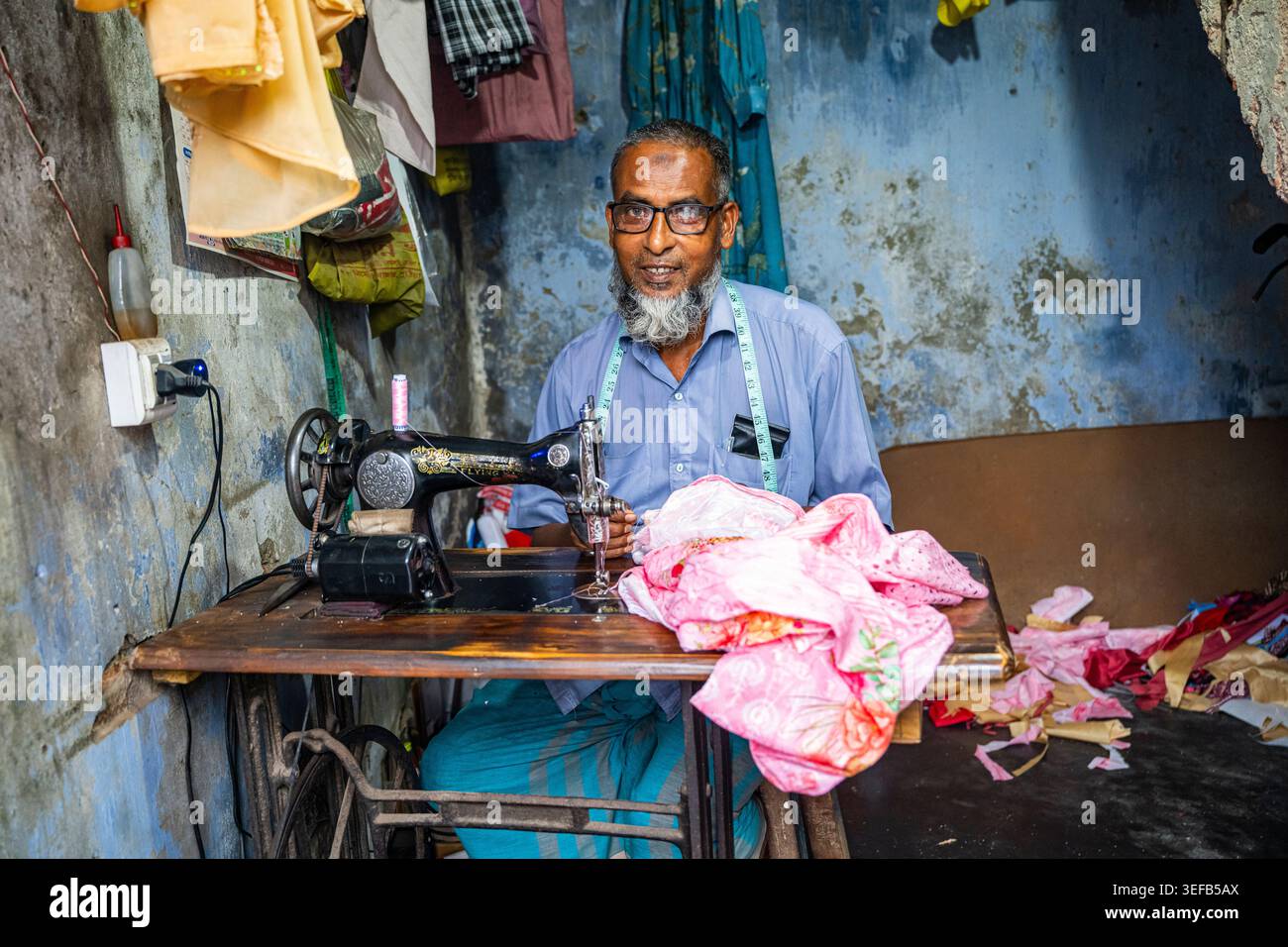 Tailleur travaillant à sa machine à coudre dans le complexe Puthia Rajbari avec des tissus colorés autour de lui au Bangladesh Banque D'Images