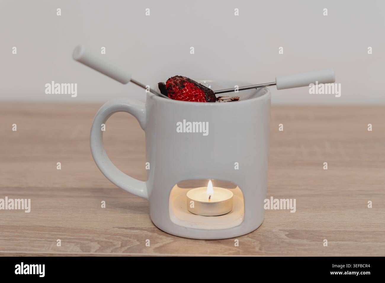 Mug à fondue en céramique blanche à la fraise trempée dans du chocolat, réchauffée par une bougie allumée, reposant sur une surface en bois clair Banque D'Images
