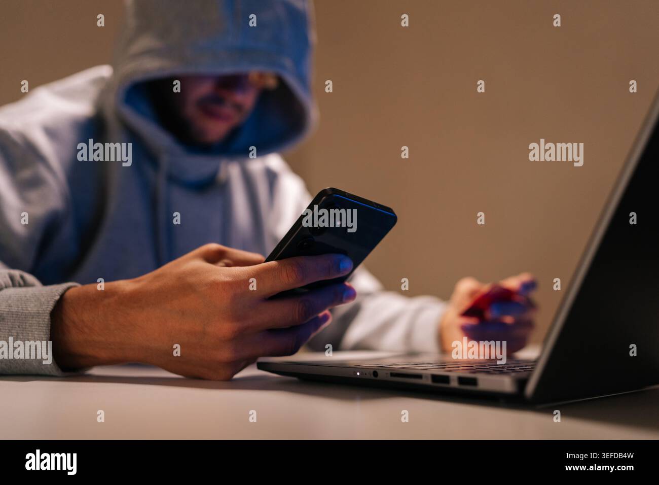 Gros plan d'une personne cagoulée se livrant à la cybercriminalité, commettant une fraude en ligne et un vol de données à l'aide d'un téléphone portable, d'un ordinateur portable et d'une carte de crédit dans l'obscurité Banque D'Images