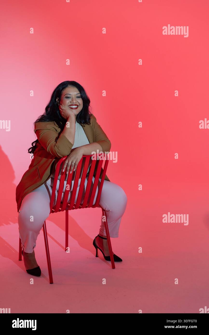 Femme adulte moyenne assise sur la chaise rouge arrière dans le studio, portant blazer tan, souriant Banque D'Images