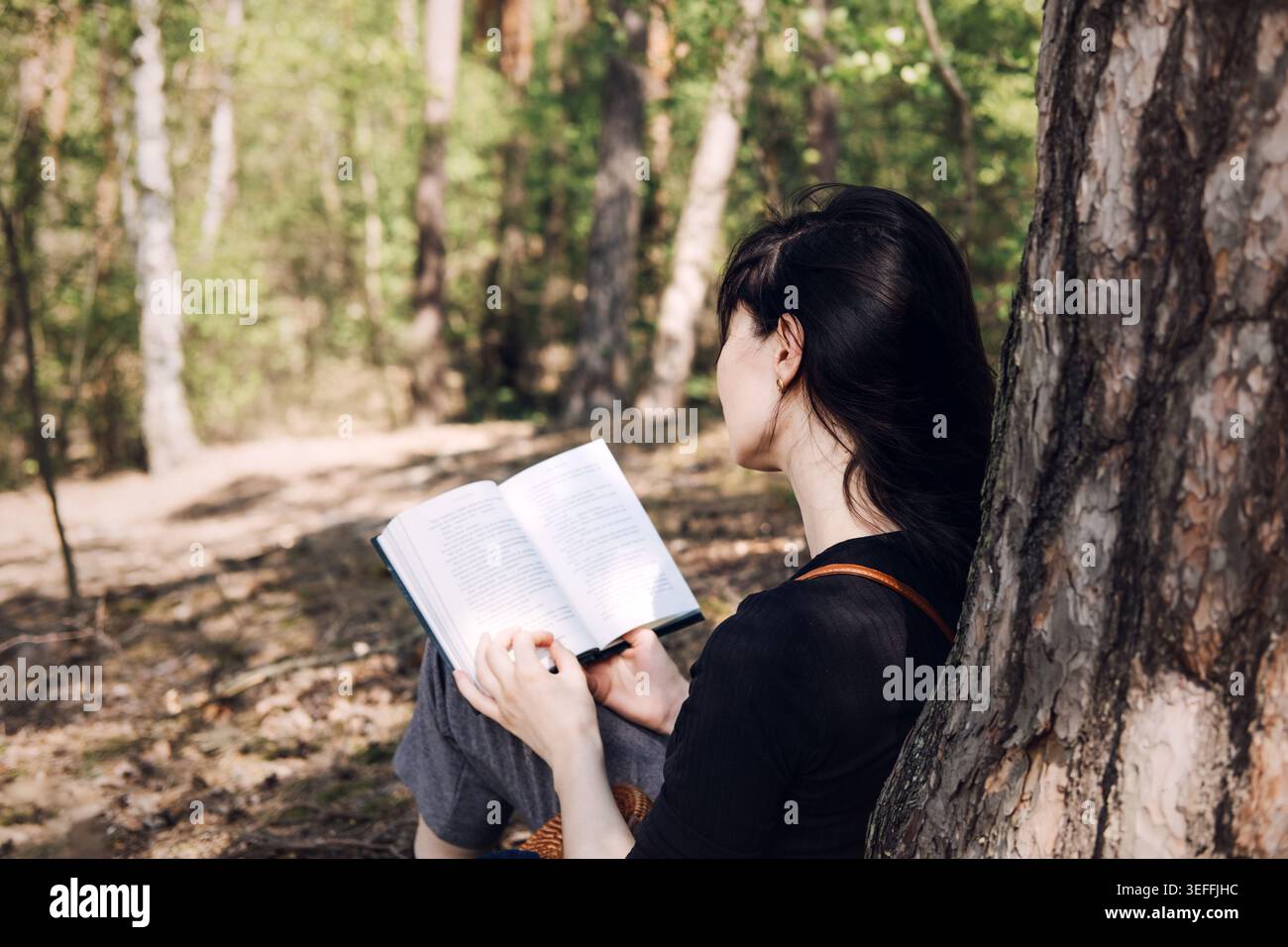 Personne s'appuie contre le tronc d'arbre tout en lisant le livre dans les bois ensoleillés. Concept de détox numérique, vie déconnectée, activité hors ligne, mouvement lent Banque D'Images