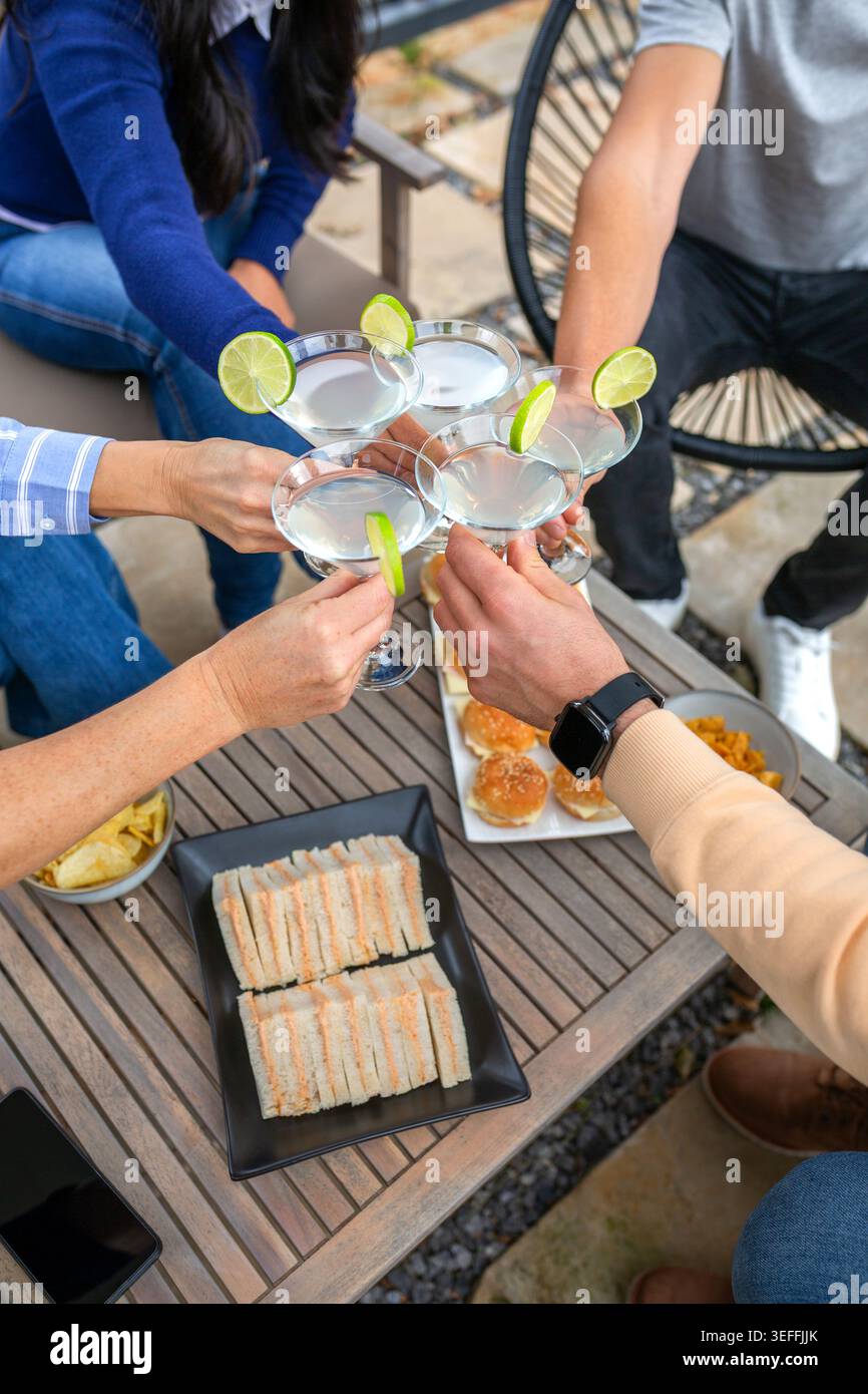 Groupe diversifié d'amis ayant du plaisir et griller des cocktails martini avec des tranches de citron vert, appréciant des collations comme des sliders et des chips de pommes de terre dans le patio extérieur, s. Banque D'Images