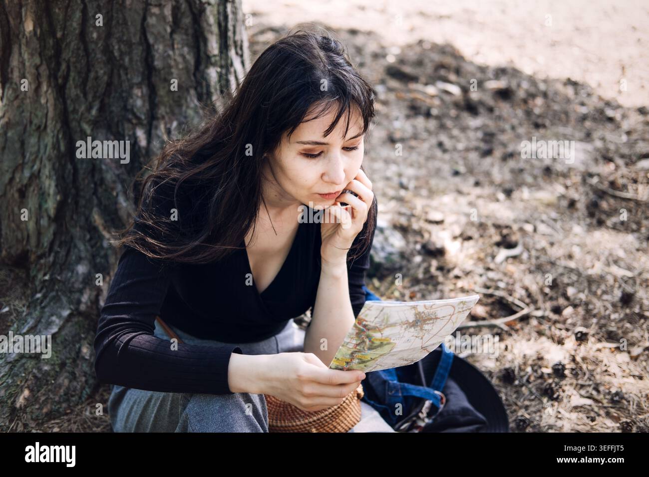 La personne est assise sous l'arbre tout en lisant attentivement la carte papier dans la forêt. Lecture de la carte papier, trouver le chemin, perdu dans le concept des bois, planification d'itinéraire Banque D'Images
