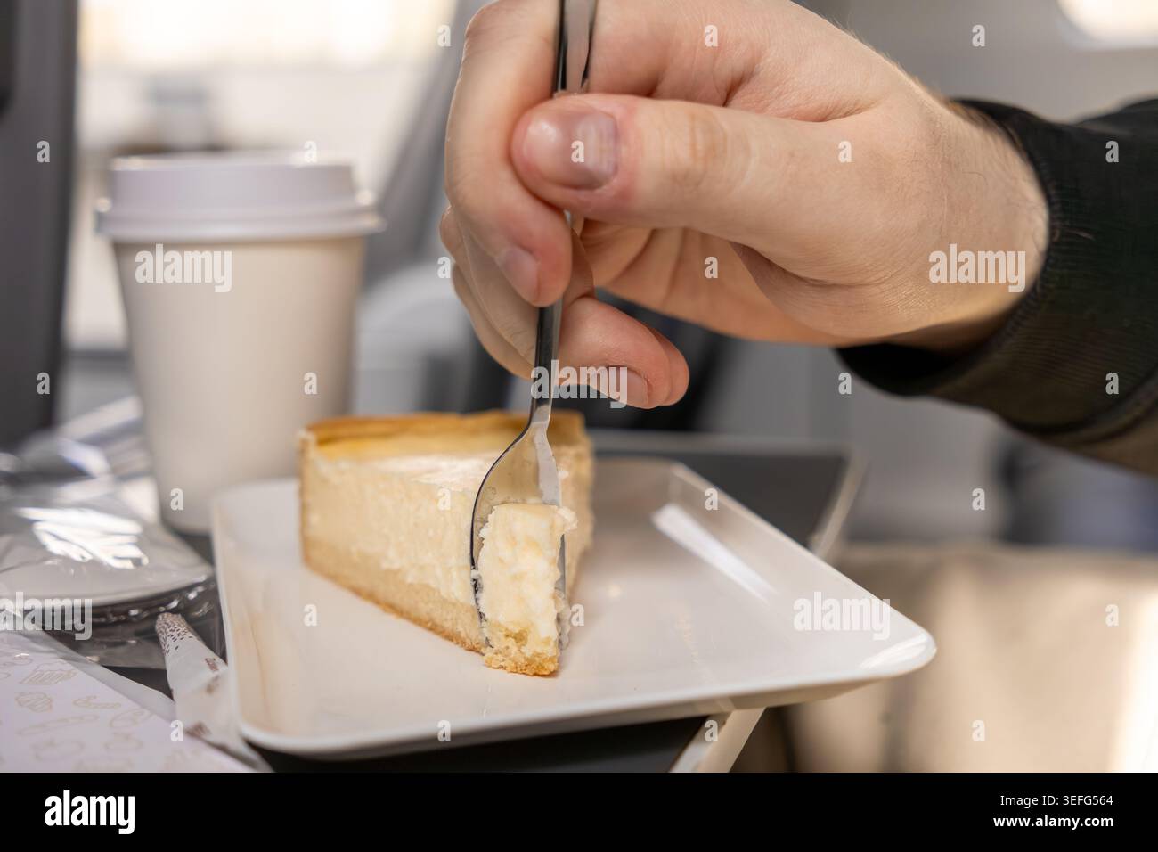 Personne mangeant une tranche de cheesecake fraîche avec une cuillère en métal sur une table de plateau d'avion à côté d'une tasse de café à emporter, capturant l'expérience du chat à bord Banque D'Images