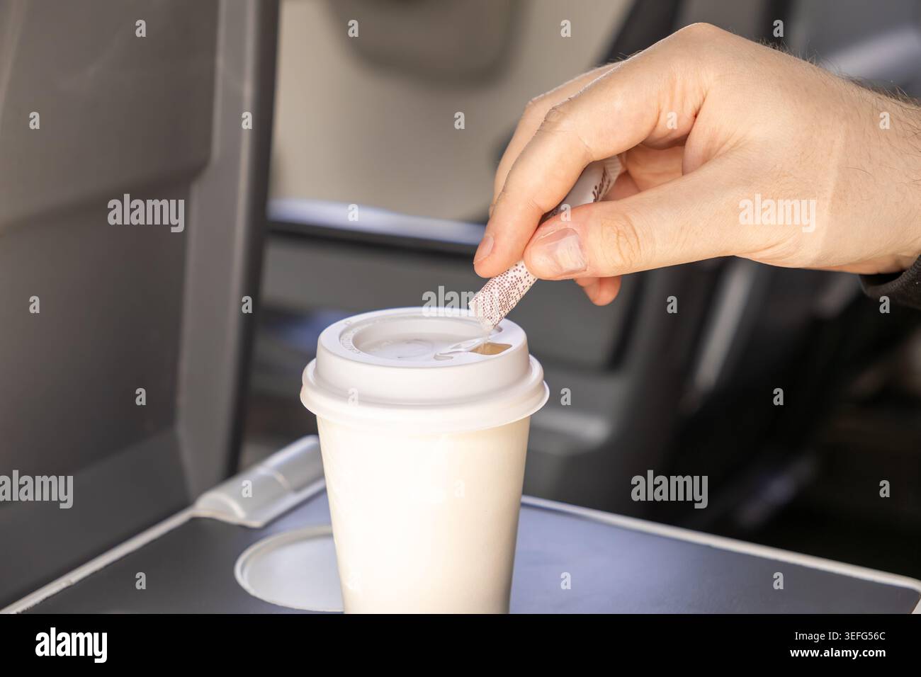 Gros plan d'une main de voyageur versant un sachet d'édulcorant dans un gobelet en papier sur un plateau d'avion pliable, représentant le confort et la culture de service à aviat Banque D'Images