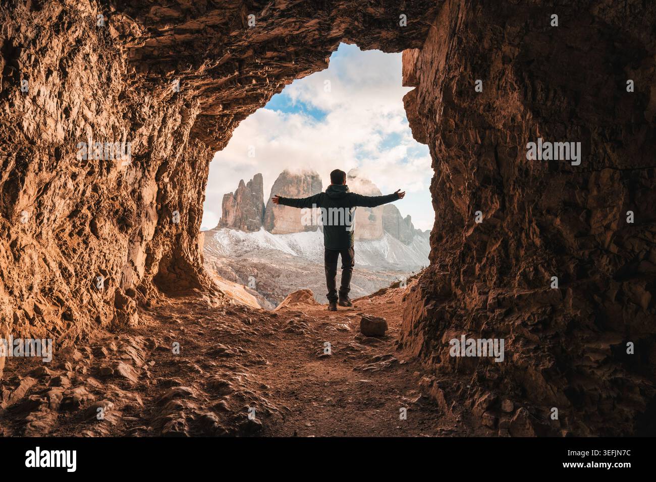 Vue arrière de l'homme debout avec les bras ouverts à l'avant de la grotte rocheuse Grotta delle Tre Cime avec pic de montagne pendant la soirée dans les Dolomites, Italie Banque D'Images