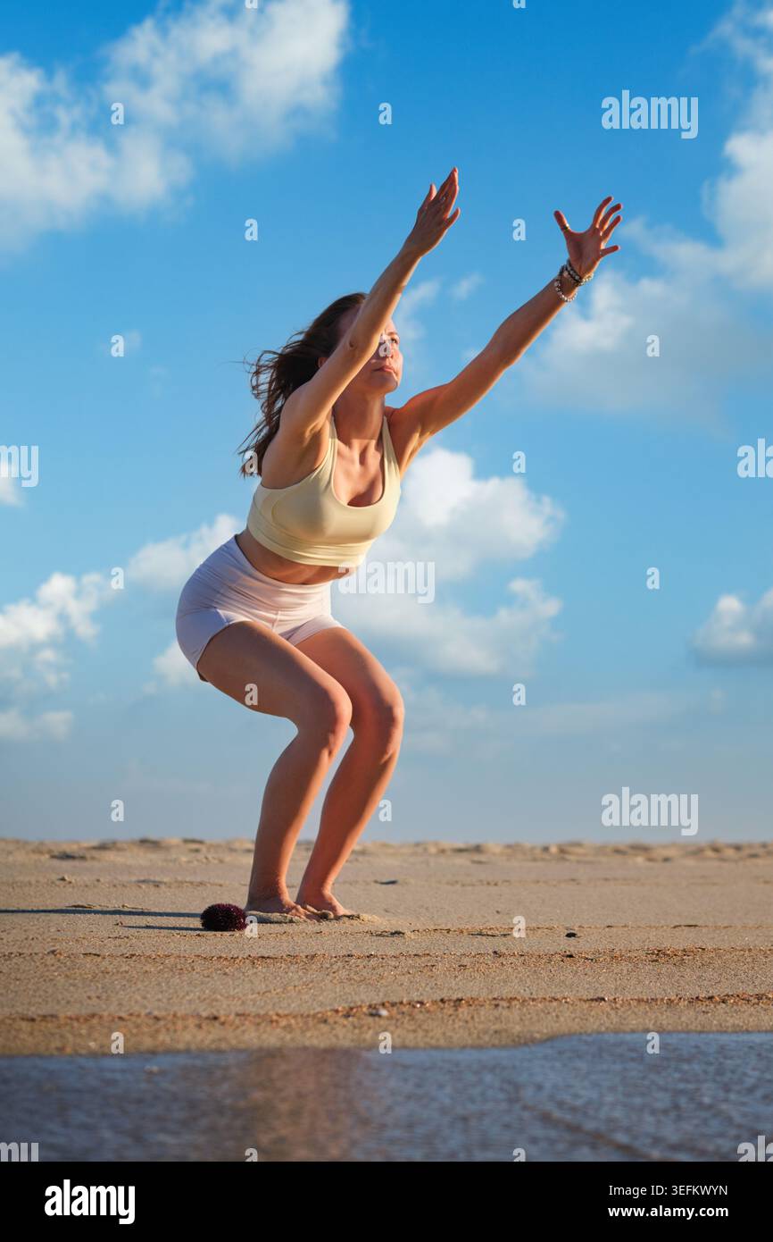 Femme pratiquant Utkatasana, ou pose de chaise, sur une plage de sable avec les bras levés, l'eau peu profonde et un ciel bleu brillant avec des nuages doux. Banque D'Images