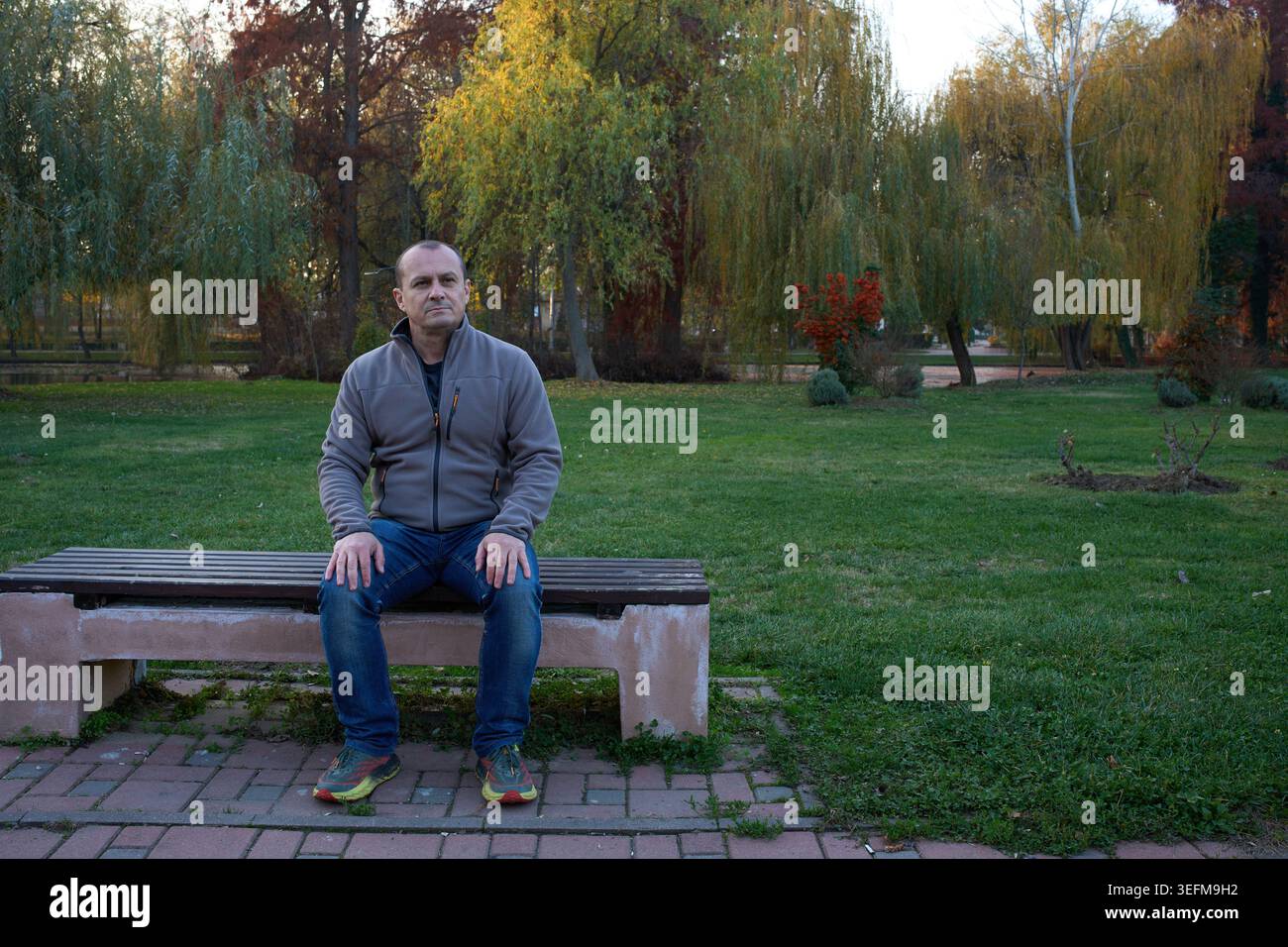 Homme d'âge moyen assis seul sur un banc de parc, portant une veste polaire grise et un Jean, regardant directement l'appareil photo avec un expert sérieux et pensif Banque D'Images