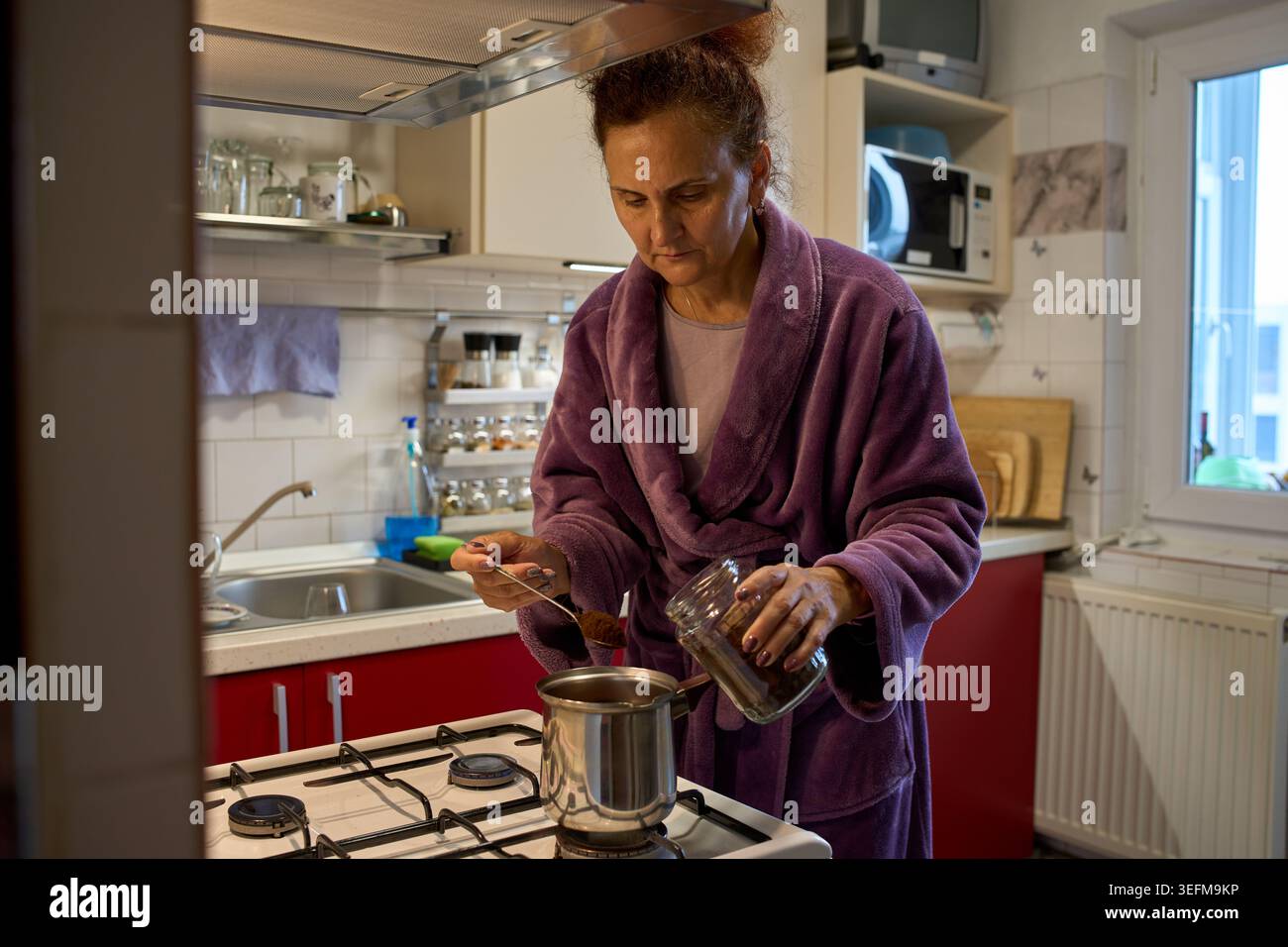 Une femme mature dans un peignoir violet cuillère du café moulu d'un pot dans un pot sur une cuisinière à gaz dans une cuisine à domicile Banque D'Images