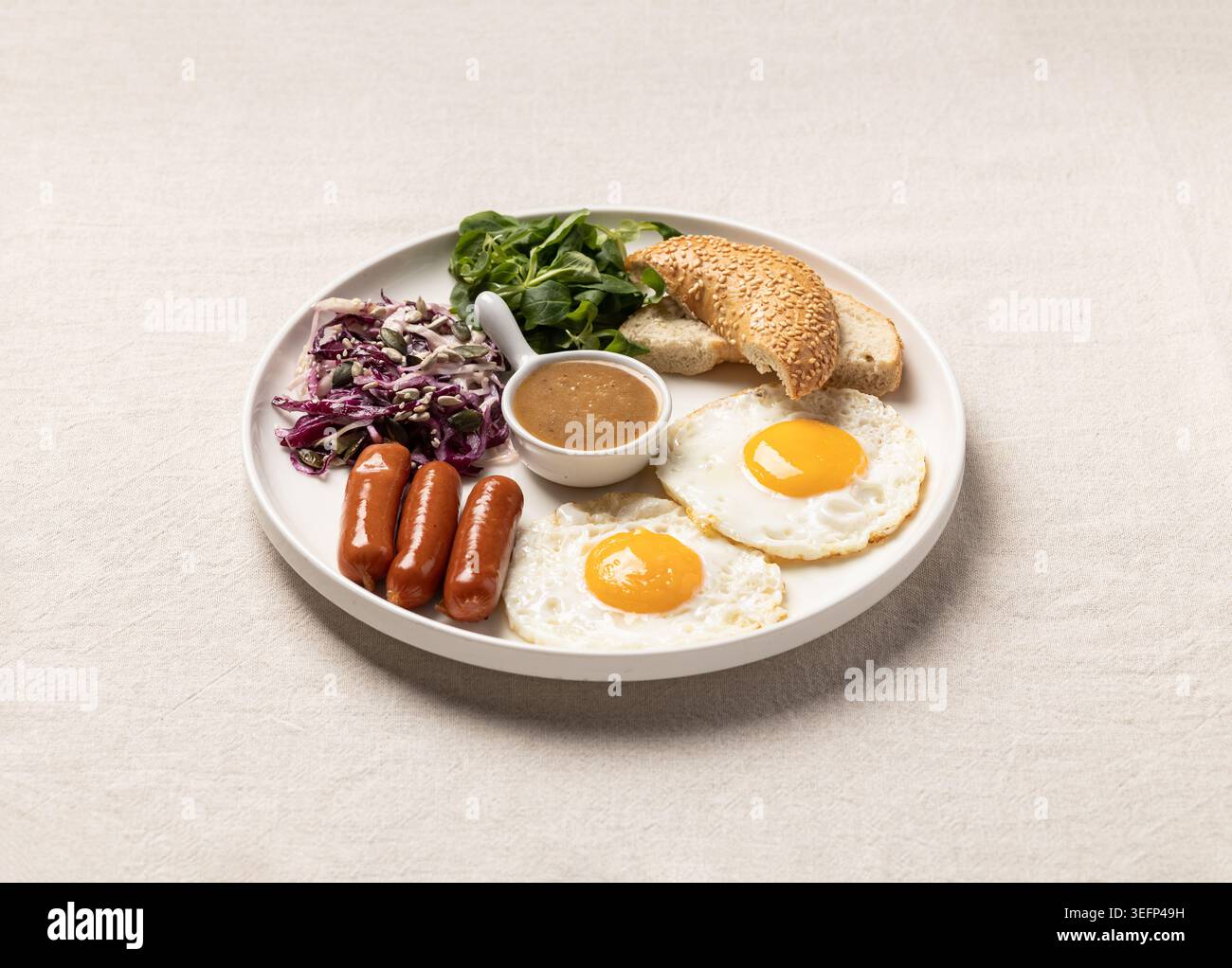 Une assiette de petit déjeuner complète avec des œufs frits, des saucisses, des légumes verts frais, du pain et de la sauce disposés soigneusement sur un plat blanc. Idéal pour les menus de restaurant, nourriture blo Banque D'Images
