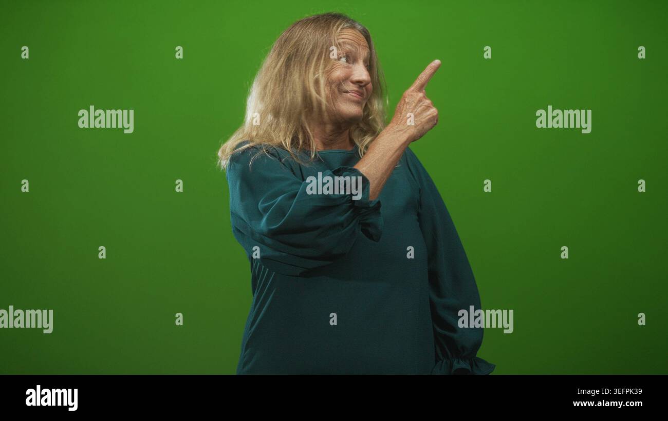 Femme senior avec la main visible pointe doigt en haut à droite tout en souriant, portant une robe sarcelle en studio ; sagesse ludique. Banque D'Images