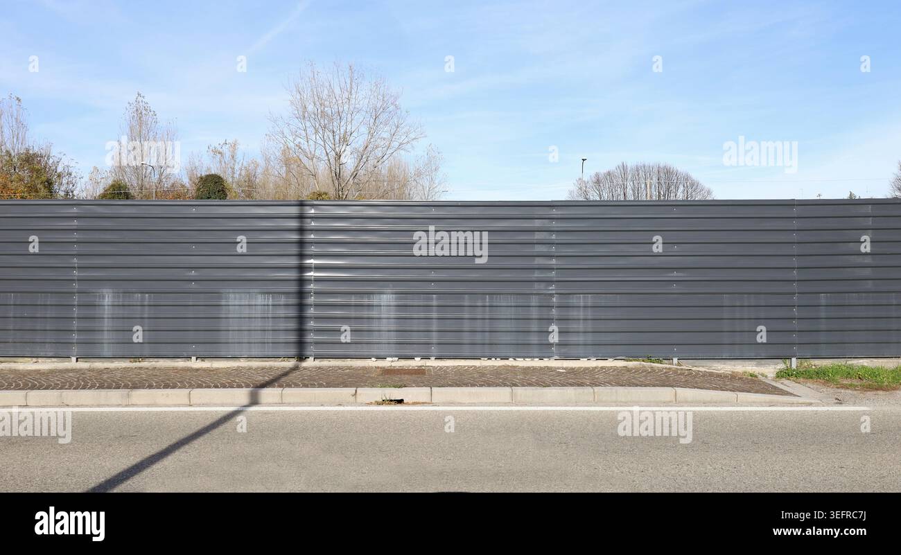 Clôture en métal ondulé noir au bord de la route. Arbres et ciel derrière, trottoir et rue devant. Arrière-plan de l'espace de copie. Banque D'Images