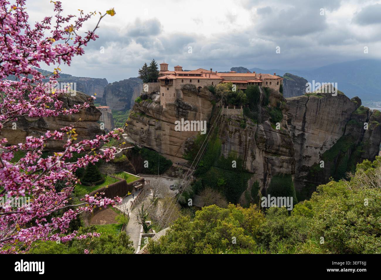 Une vue spectaculaire sur le monastère Saint de Varlaam encadré par un arbre Judas en fleurs, sur fond d'anciens piliers rocheux et d'un ciel nuageux. Banque D'Images