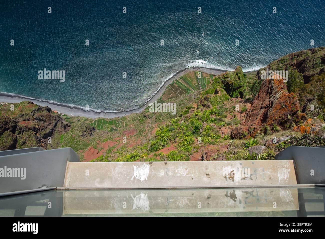Funchal, Madère, 12 mai 2026. Vue d'en haut montrant l'océan bleu profond, une plage rocheuse et des champs en terrasses à la base de falaises vertes et rouges abruptes Banque D'Images