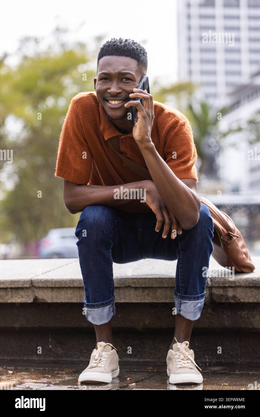 Homme accroupi sur le rebord de pierre par l'eau à plaza, tenant le téléphone, polo rouille, sac à dos à côté Banque D'Images