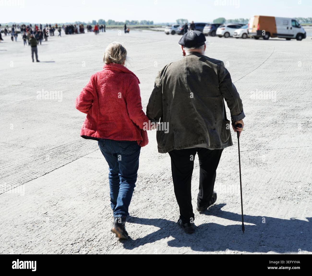 Un couple de personnes âgées marche main dans la main à travers un parking en béton. La femme porte une veste rouge vif et un Jean bleu, tandis que l'homme porte un manteau foncé, Banque D'Images