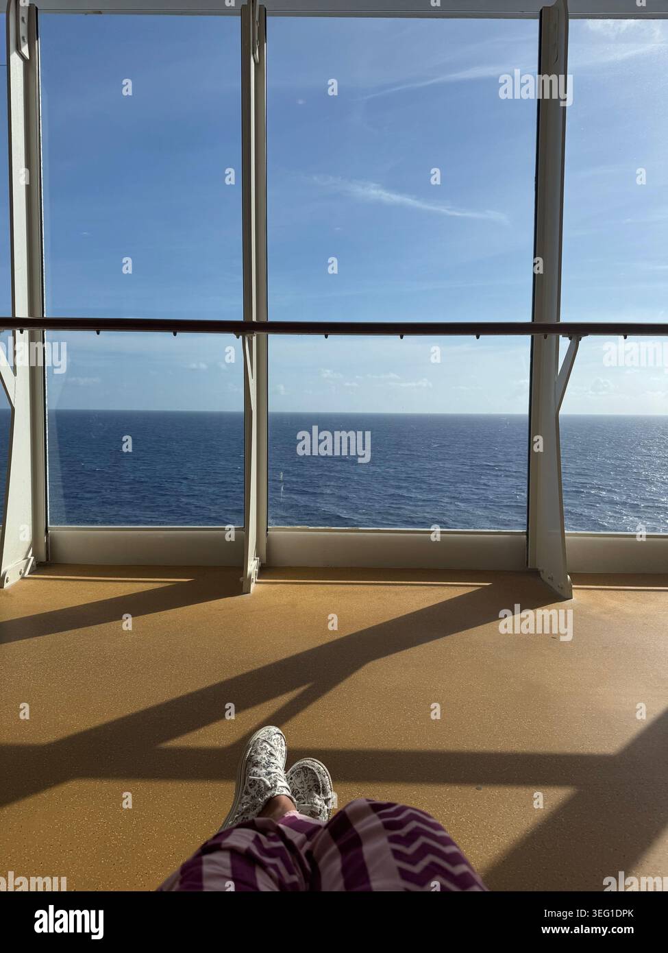 Vue à la première personne des pieds d'un voyageur reposant sur une terrasse couverte ensoleillée près d'une immense fenêtre panoramique donnant sur l'horizon calme de l'océan. - Image de stock capturée avec un smartphone