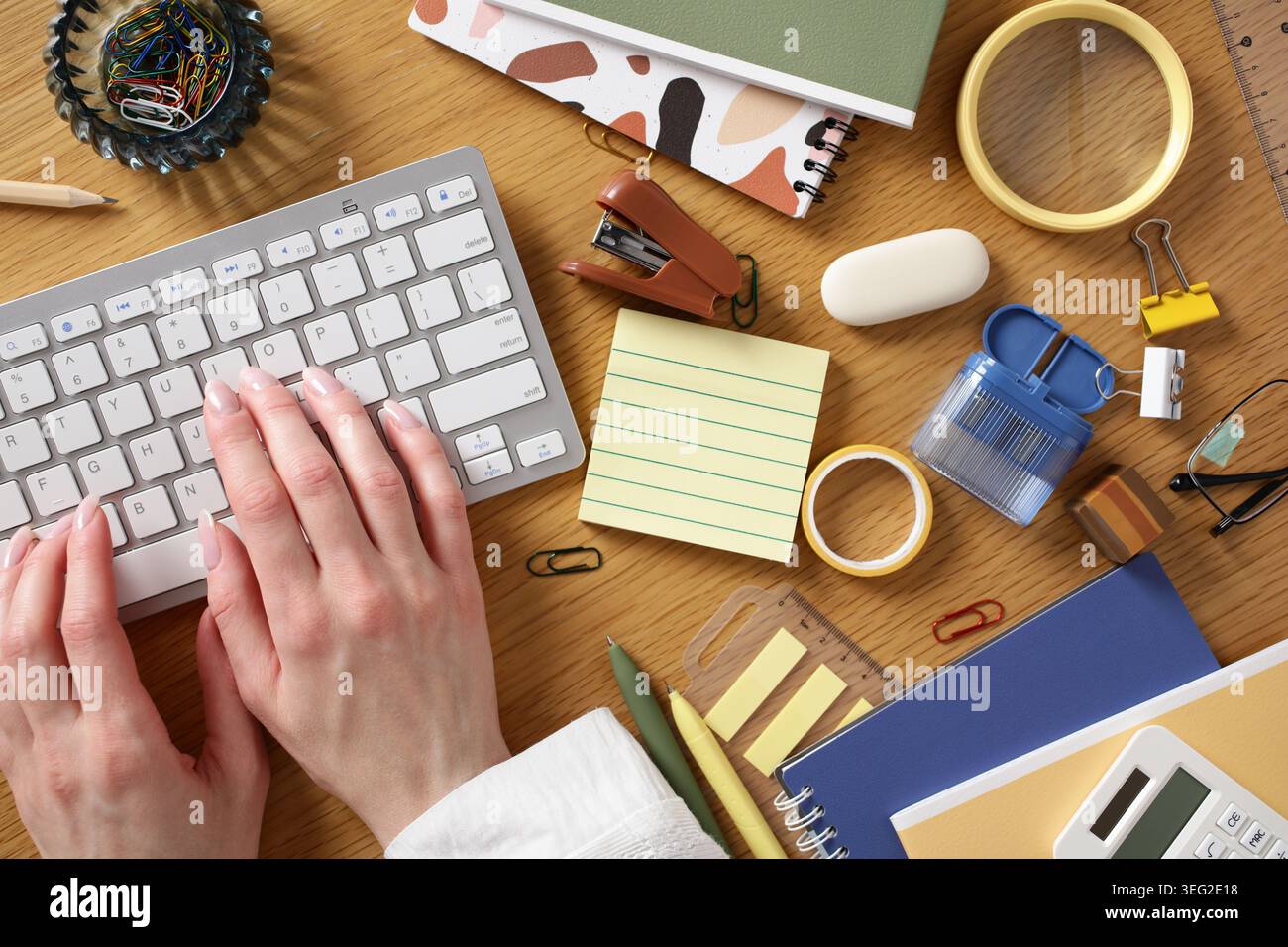 Vue de dessus d'un bureau à domicile contemporain posé à plat avec les mains tapant sur un clavier, des notes adhésives, un carnet de notes, du ruban adhésif, des clips, et autre papeterie pour le produit Banque D'Images