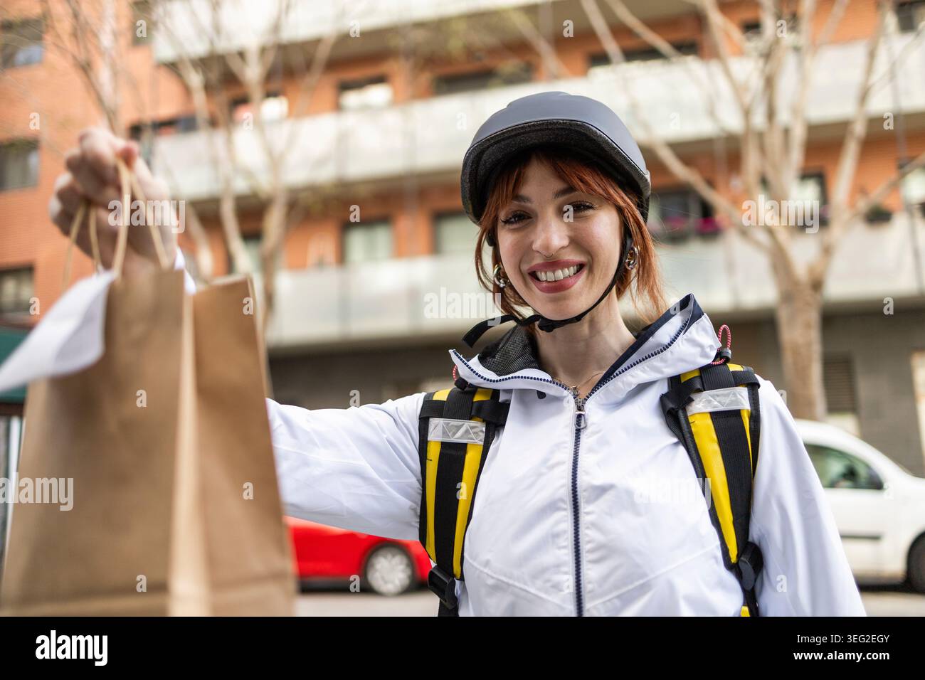 Femme souriante livreur portant un casque et une veste remet un sac en papier à emporter au client à l'extérieur dans une rue de la ville, mettant en vedette la livraison de nourriture rapide Banque D'Images