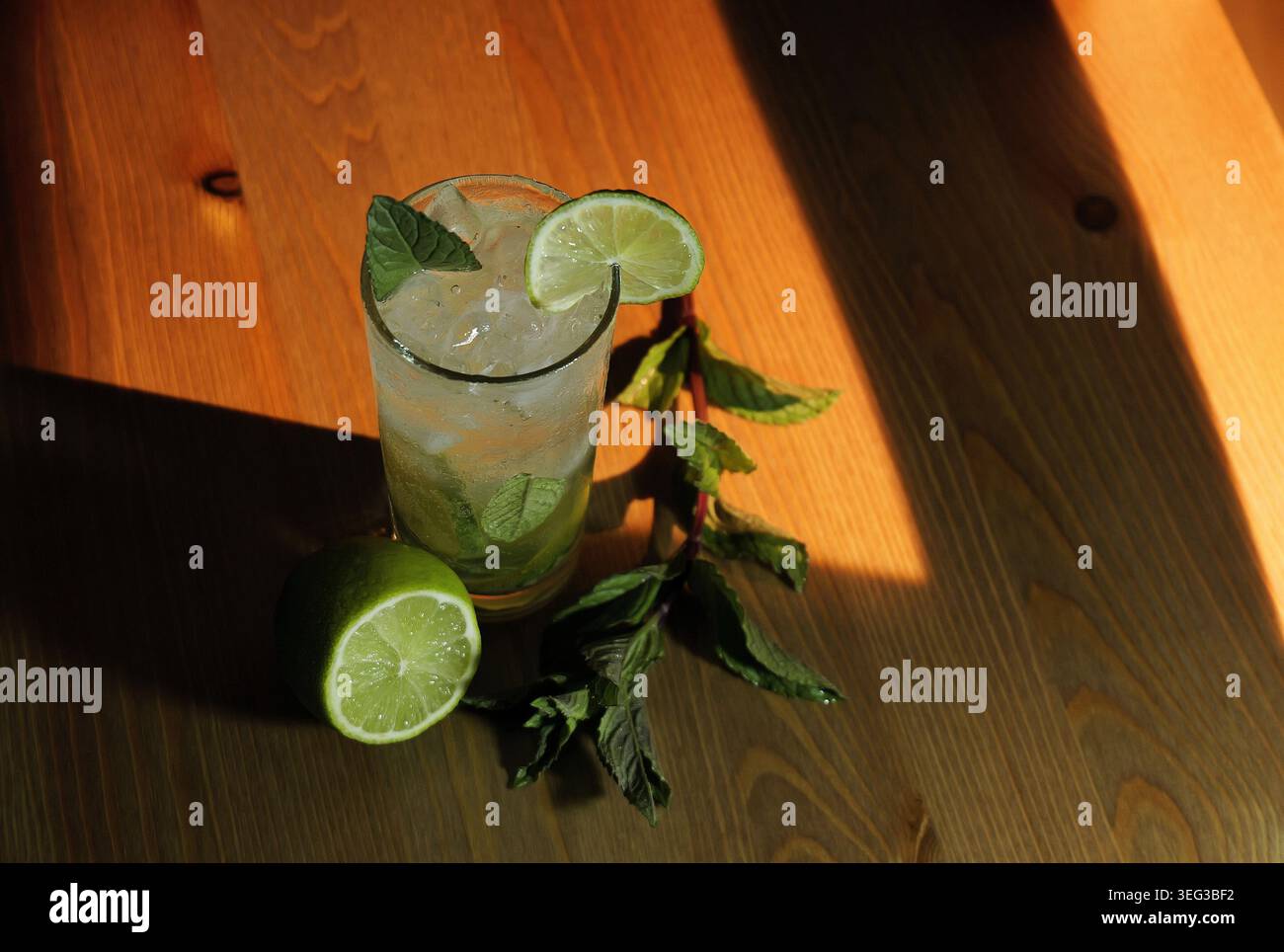 Cocktail de mojito tenant de la glace, des tranches de citron vert et des feuilles de menthe fraîche dans un grand verre, assis sur une surface en bois avec une branche entière de citron vert et de menthe, de l'illumin Banque D'Images