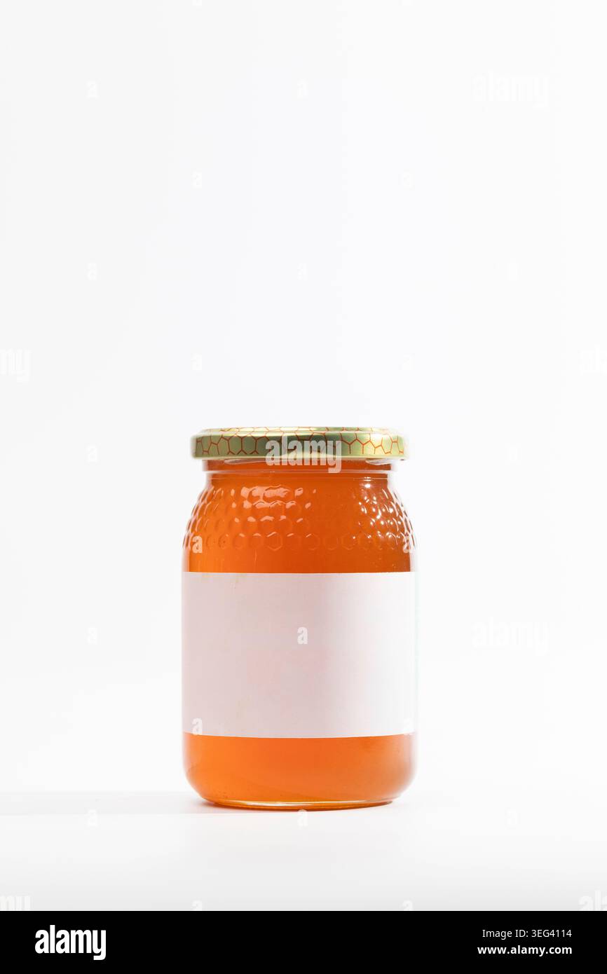 Le pot en verre transparent contient du miel naturel doré de fleurs sauvages. Le couvercle en métal ferme hermétiquement le bocal. L'étiquette de maquette s'enroule autour du pot. Le miel scintille sous brillant Banque D'Images