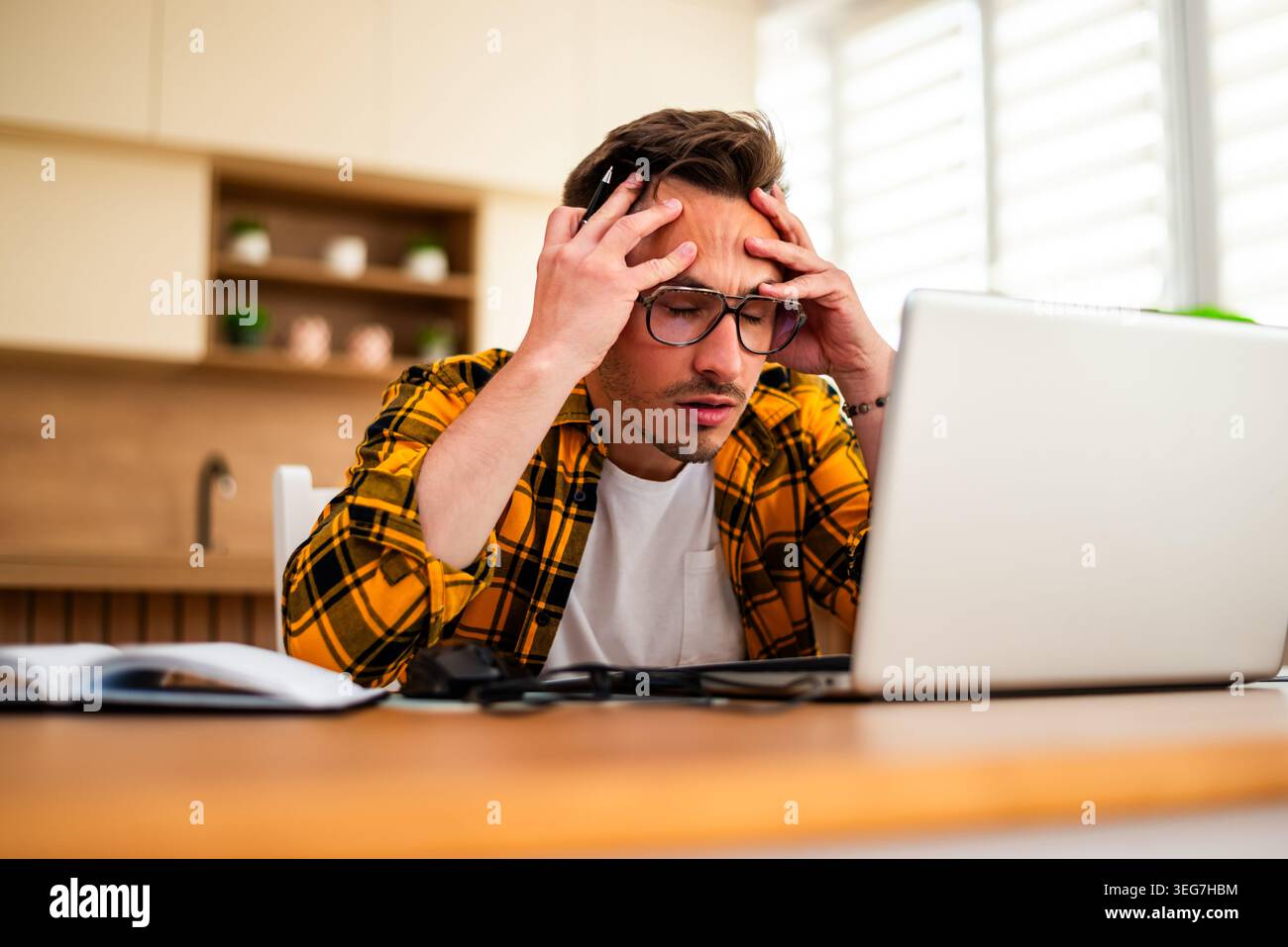 Un jeune homme stressé travaille sur son ordinateur portable tout en tenant un stylo, en posant sa tête dans sa main, montrant des signes de frustration ou de fatigue mentale. Banque D'Images