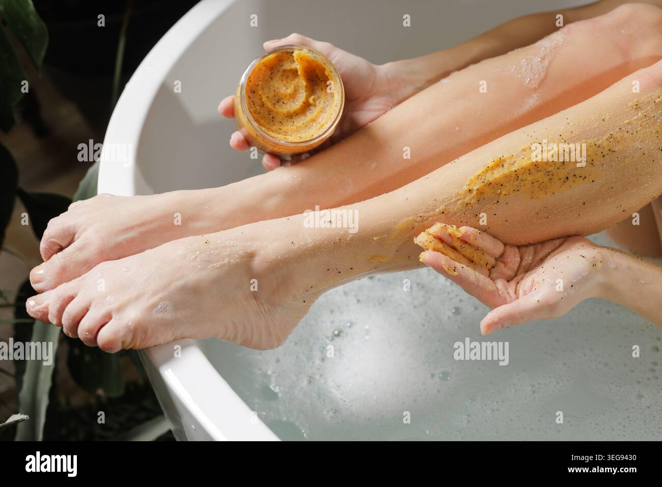 Une femme applique un gommage corporel naturel sur ses jambes tout en se relaxant dans une baignoire, favorisant une peau saine et des routines de soins personnels. Banque D'Images