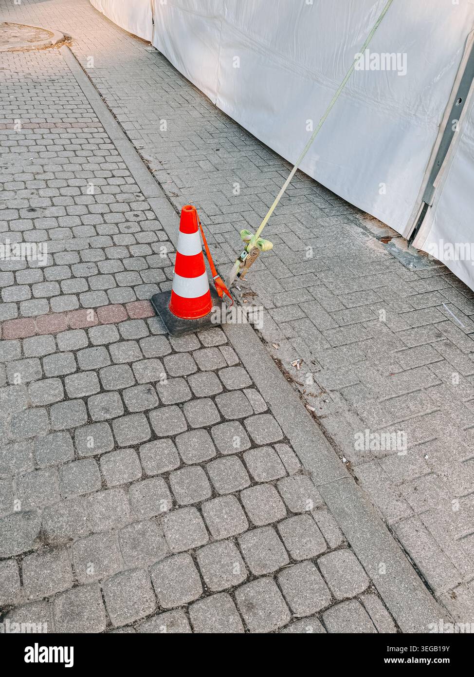 Cône de signalisation orange marquant une sangle de tension attachée à une structure extérieure temporaire sur un trottoir pavé de la ville. Scène de sécurité urbaine avec construction, Banque D'Images