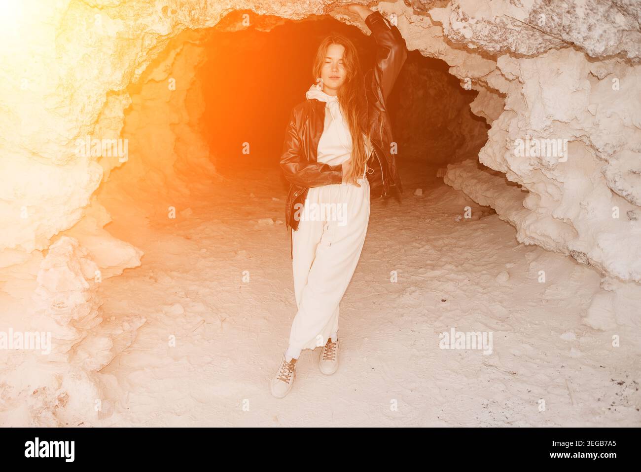 Femme caverne nature posant dans une caverne rocheuse avec une lumière chaude du soleil tout en portant une veste en cuir élégante et un survêtement blanc Banque D'Images