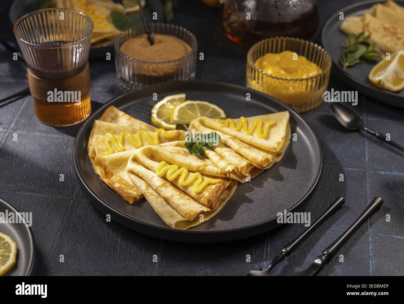 Fines crêpes pliées sur une assiette noire, garnies de caillé de citron brillant, de menthe fraîche et d'une tranche de citron, servies avec du sucre, du thé chaud et du fromage blanc supplémentaire pour Banque D'Images