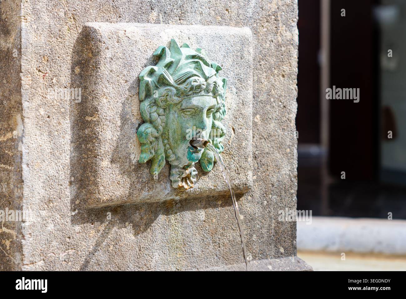 Gros plan d'une fontaine murale en pierre altérée avec un lion patine vert ou un bec verseur mythique, l'eau ruisselant de la bouche à la lumière du jour. Banque D'Images
