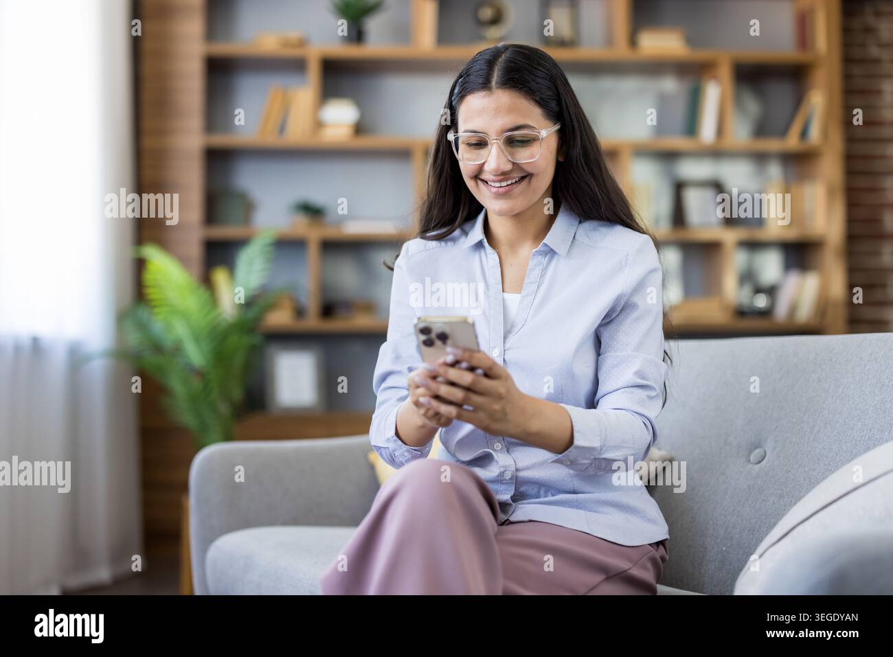 Femme portant des lunettes assise sur un canapé confortable dans un salon moderne, souriant joyeusement tout en envoyant des SMS sur son téléphone portable, en se connectant et en communiquant en ligne avec la technologie Banque D'Images