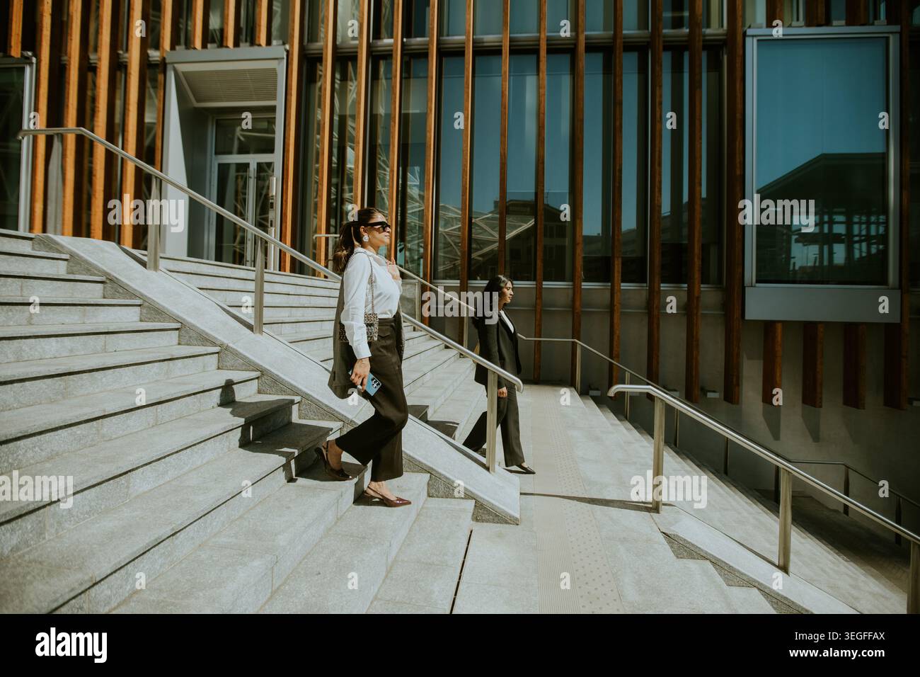 Deux professionnels marchent sur les marches d'un bâtiment moderne par un après-midi ensoleillé. Une personne est habillée en blanc avec des lunettes de soleil, tandis que Banque D'Images