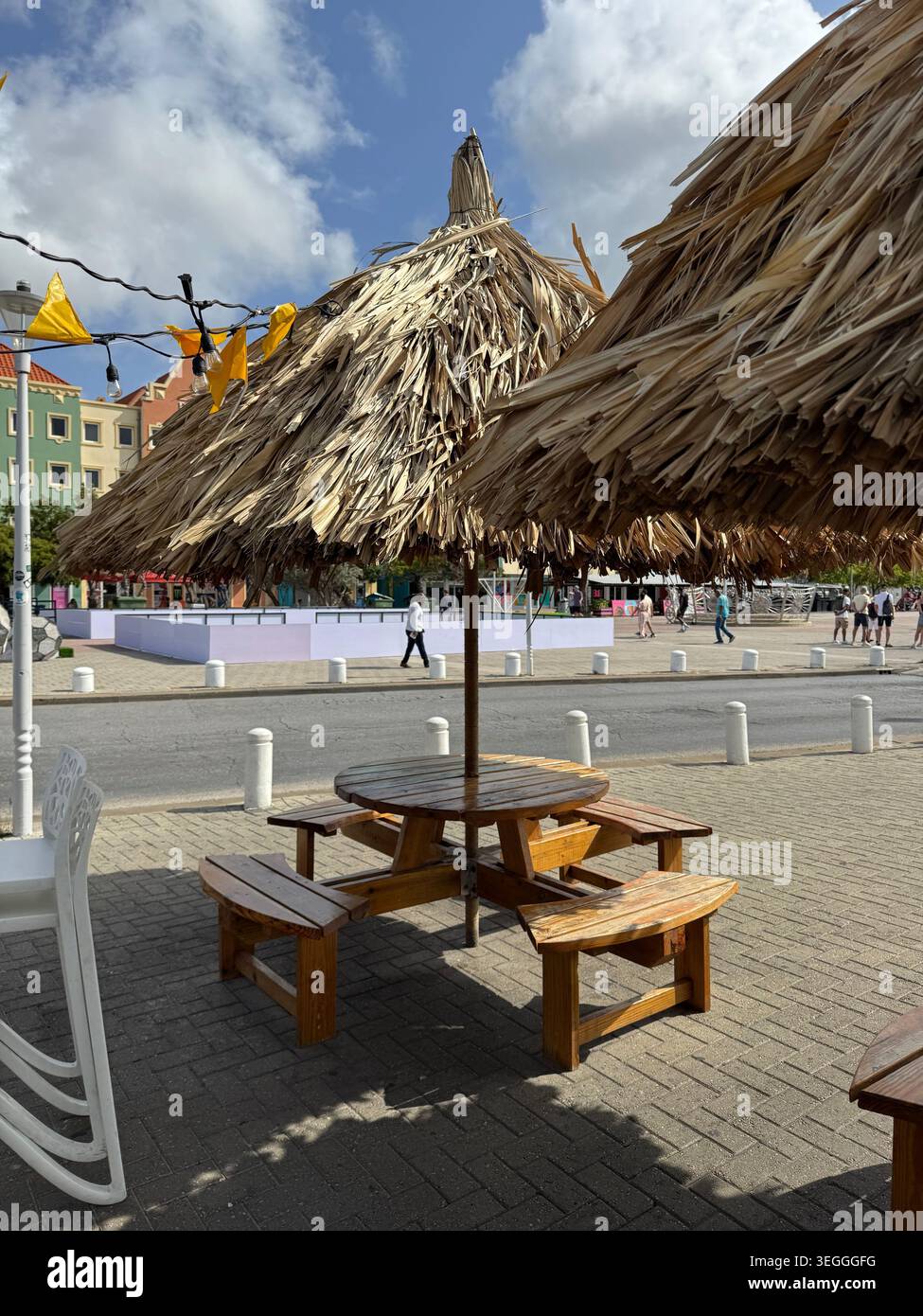 Une terrasse de café en plein air avec des tables en bois et un grand parasol en paille de chaume dans une place publique de Willemstad, Curaçao. - Image de stock capturée avec un smartphone