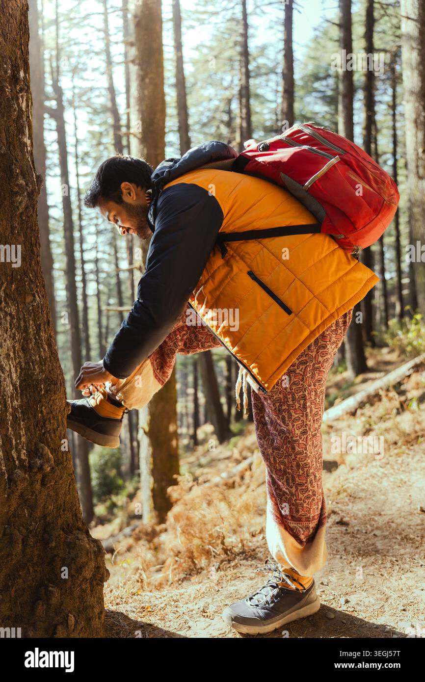 Un jeune homme portant un gilet jaune et un sac à dos rouge attache ses bottes de randonnée sur un chemin de terre dans une forêt de pins à Chakrata, Uttarakhand, Inde. Banque D'Images