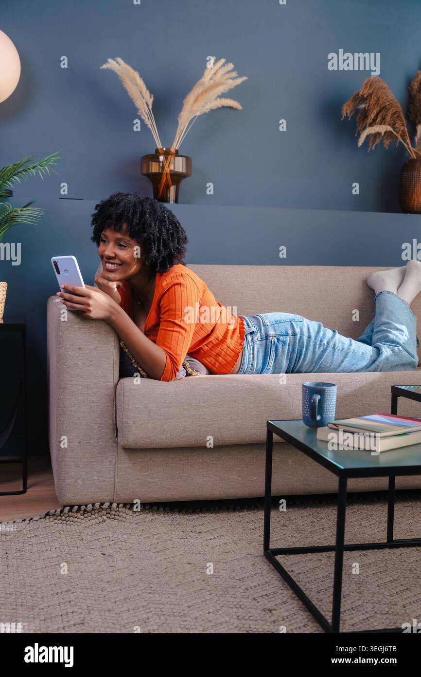 Portrait vertical femme afro-américaine allongée sur un canapé et utilisant son smartphone à la maison Banque D'Images