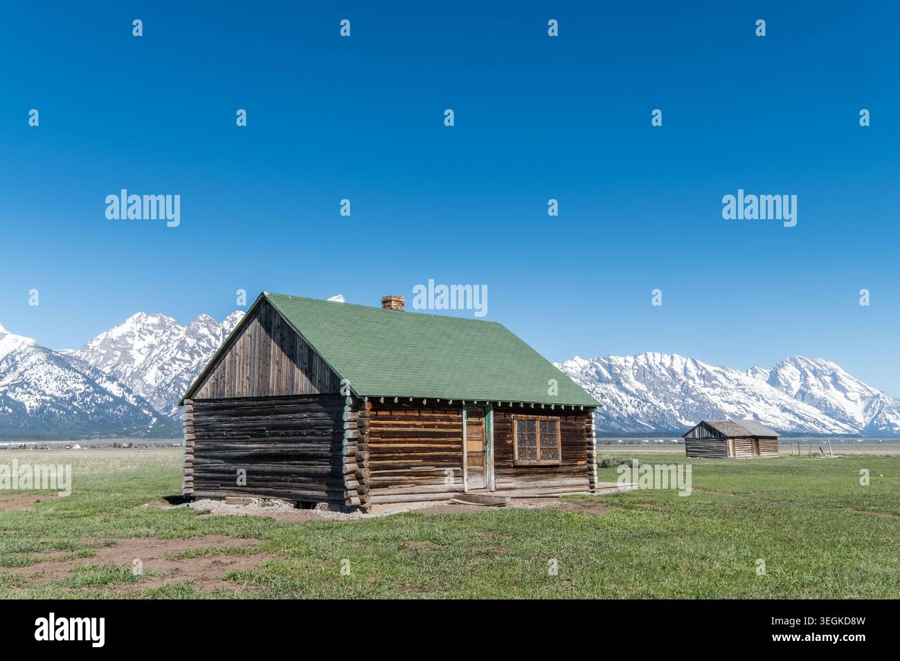 Cabane historique en rondins dans un vaste champ verdoyant avec de majestueuses montagnes enneigées sous un ciel bleu clair. Serein et pittoresque. Idéal pour les voyages, la nature, Banque D'Images