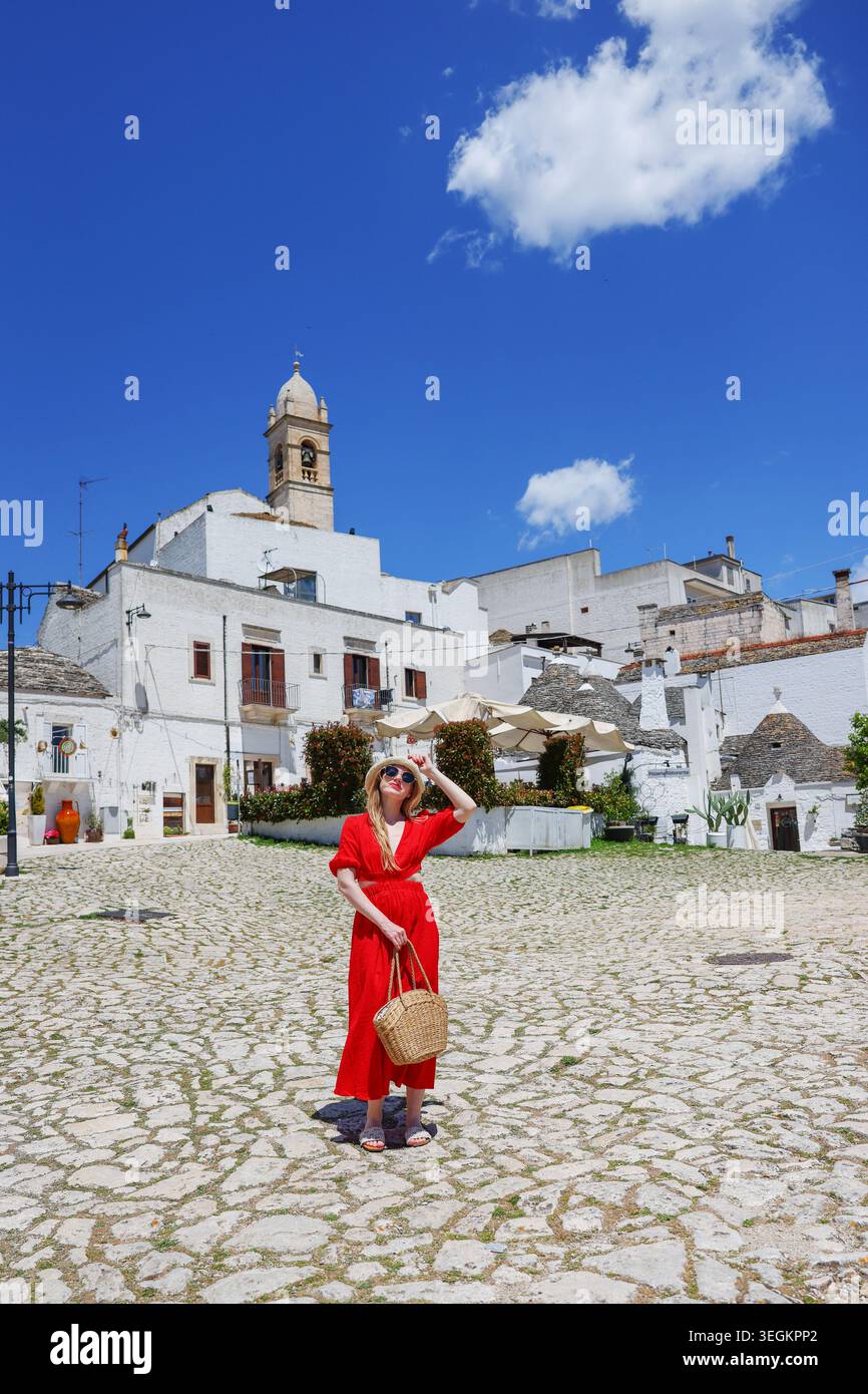 Femme élégante en robe rouge marchant à travers la place ensoleillée à Alberobello, Italie avec des maisons de trulli emblématiques et des vibrations estivales méditerranéennes. Banque D'Images